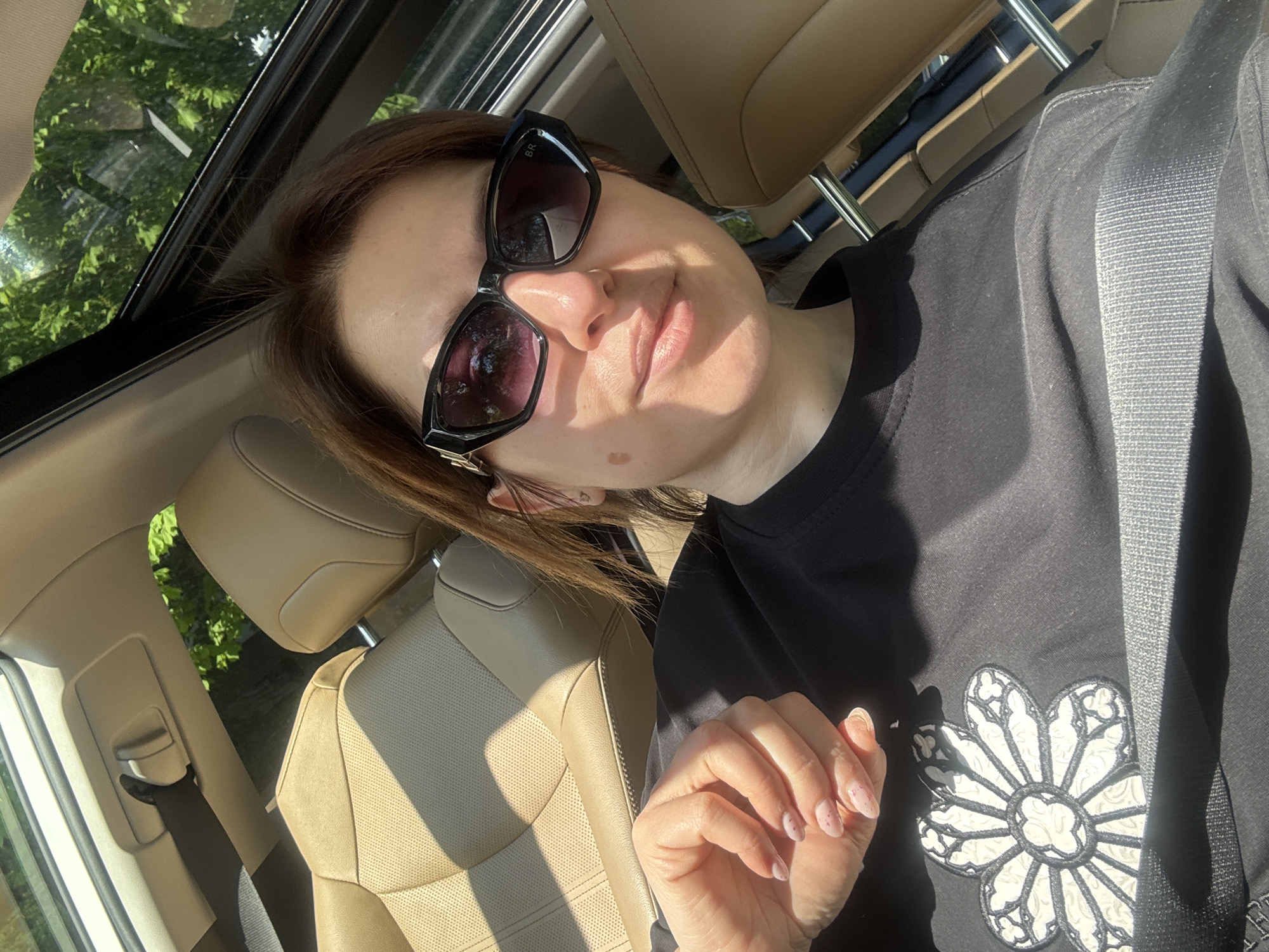 The image shows a woman in a car, likely a passenger, wearing sunglasses and a black sweatshirt with a white flower design. The car appears to be in motion. Light streams through a sunroof, illuminating the beige leather interior. The background shows green foliage, suggesting the car is near trees or a park. The photo's location within Sofia, Bulgaria, cannot be definitively determined from the provided visual information.