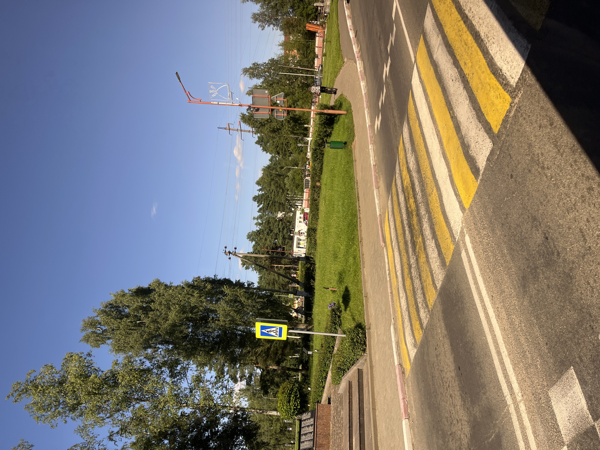 The image depicts a street scene in Pastavy, Belarus, on a clear, sunny day. In the foreground, a well-marked crosswalk with alternating yellow and white stripes dominates the asphalt road. The sidewalk adjacent to the crosswalk features a pink and white striped border. To the left of the crosswalk, a monument with steps and a granite plaque is partially visible. A pedestrian crossing sign with a blue background and a white pedestrian symbol is positioned on a pole on the sidewalk.
The background reveals a well-maintained grassy area bordered by a low green hedge. Several tall birch trees with green foliage are scattered across the landscape, along with other trees characteristic of a park or urban green space. Beyond the trees, utility poles with wires are visible, indicating the presence of infrastructure. A red and white lamppost stands near a curved paved path. A person on a bicycle is seen riding along this path, heading away from the viewer. Further in the distance, buildings and more trees can be observed. The sky is a vibrant, clear blue with a few wispy clouds.