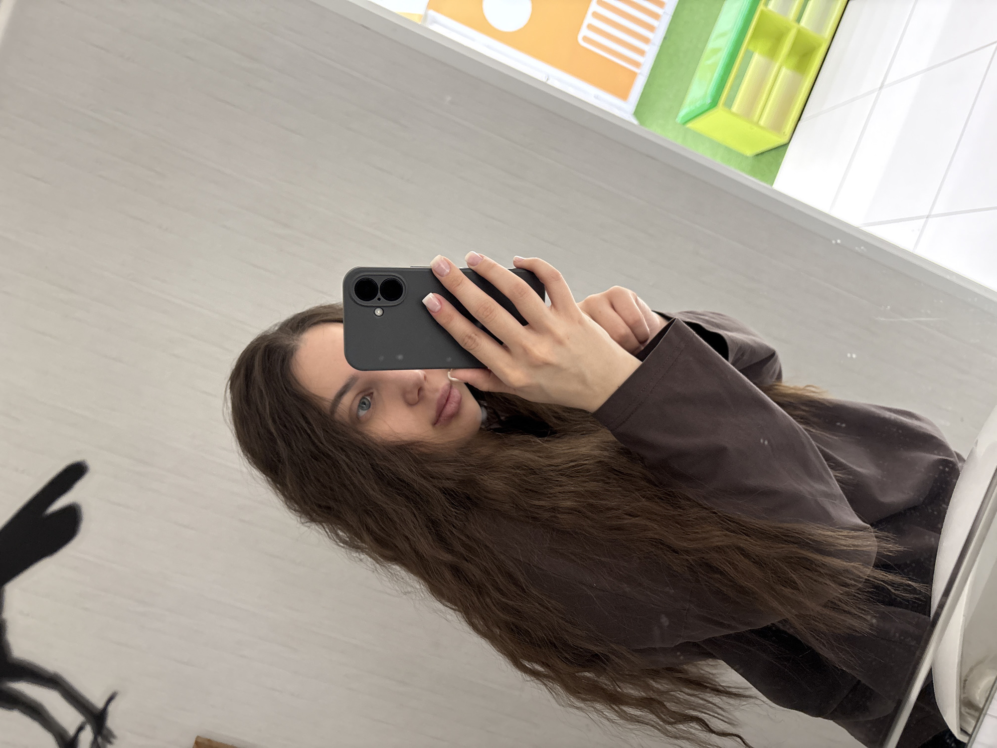 A young woman with long, wavy brown hair is taking a selfie in a bathroom mirror. She is holding a dark-colored smartphone up to her face with both hands. Her fingernails are manicured with a French tip design. She is wearing a brown top. In the background, a wall with white tiles and a colorful design is visible. A sink is partially visible on the right side of the image. A shadow of what may be a toy figure is visible in the lower left corner of the reflection. The location is presumably Chaikovsky, Russia.