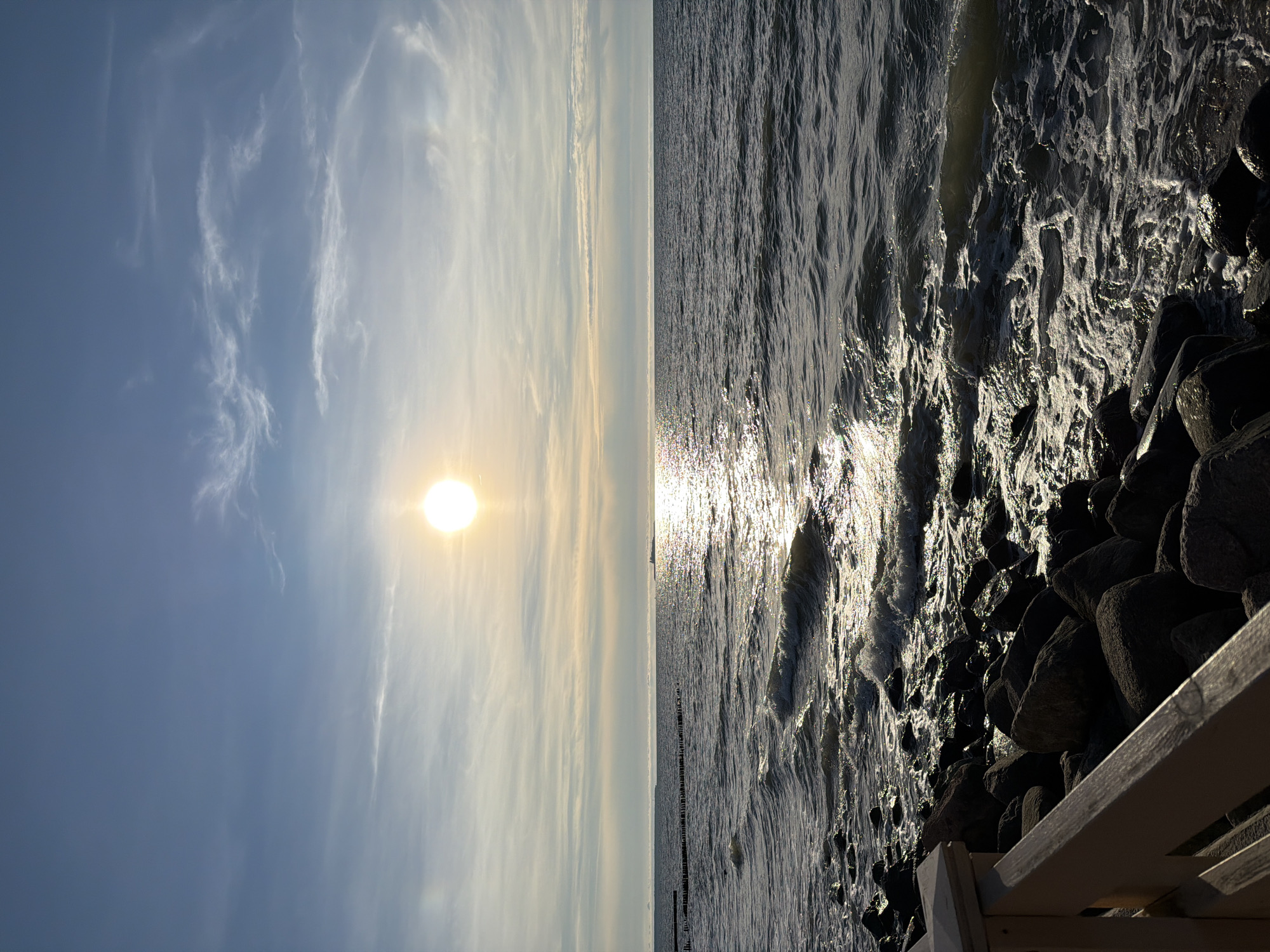 The image depicts a serene seascape with a bright sun low in the sky, casting a shimmering reflection on the water. The foreground features a rocky shoreline and a wooden railing, suggesting a coastal path or boardwalk. In the distance, a faint line of wooden posts breaks the surface of the water, possibly serving as a breakwater or marker. The sky is a gradient of blue, with wispy clouds scattered across it, indicating fair weather. The overall scene is tranquil and evokes a sense of calm. The location is Zelenogradsk, Russia.