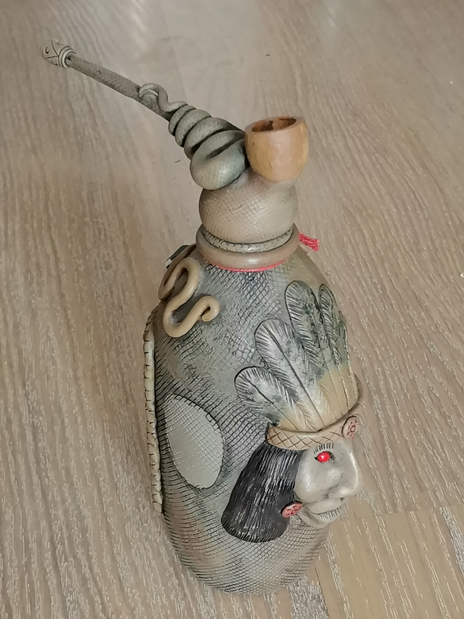 The image displays a single object, a decorative bottle, lying on a light-colored wooden surface. The bottle is elaborately designed, resembling a stylized Native American figure. It features a textured, snake-like pattern across its body and is adorned with appliqué elements such as feathers, a face with red eyes, and a coiled snake motif. A wooden pipe bowl is attached to the top, and a long, segmented tube extends upwards and to the left, also resembling a snake. A thin red cord is visible around the neck of the bottle. The lighting suggests an indoor environment. There is no information or visual indication about the city of Oryol, Russia, in this image.