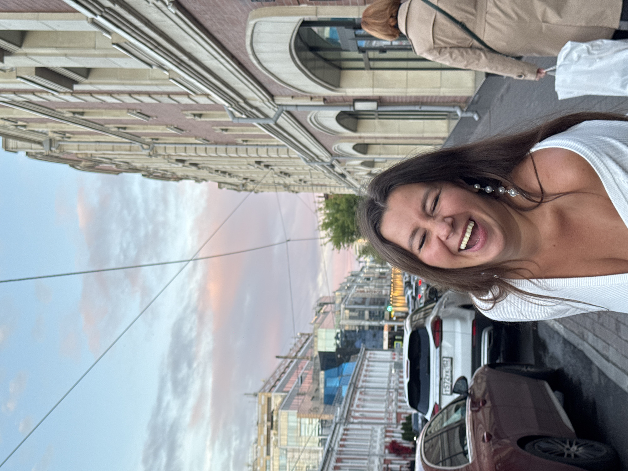 An outdoor, eye-level, medium-full shot captures a woman laughing vibrantly while standing on a sidewalk with cars parked on the street and a building lining the right side of the frame. The sky behind her is a soft mix of blue and pink clouds, suggesting either sunrise or sunset. She has long, dark brown hair and is wearing a white off-the-shoulder top and pearl earrings. Her mouth is wide open in laughter, her eyes are squinted shut, and she is looking slightly to her left. In the background, a woman in a beige trench coat is walking away from the camera, carrying a white shopping bag. The street is lined with parked cars, and the buildings on the right have an architectural style with arches and ornate detailing. The buildings on the left are a mix of older, traditional architecture and modern structures. The presence of a license plate on a white SUV that reads "F572XK 150" and the distinct architecture of the buildings suggest the location is in Nizhny Novgorod, Russia.