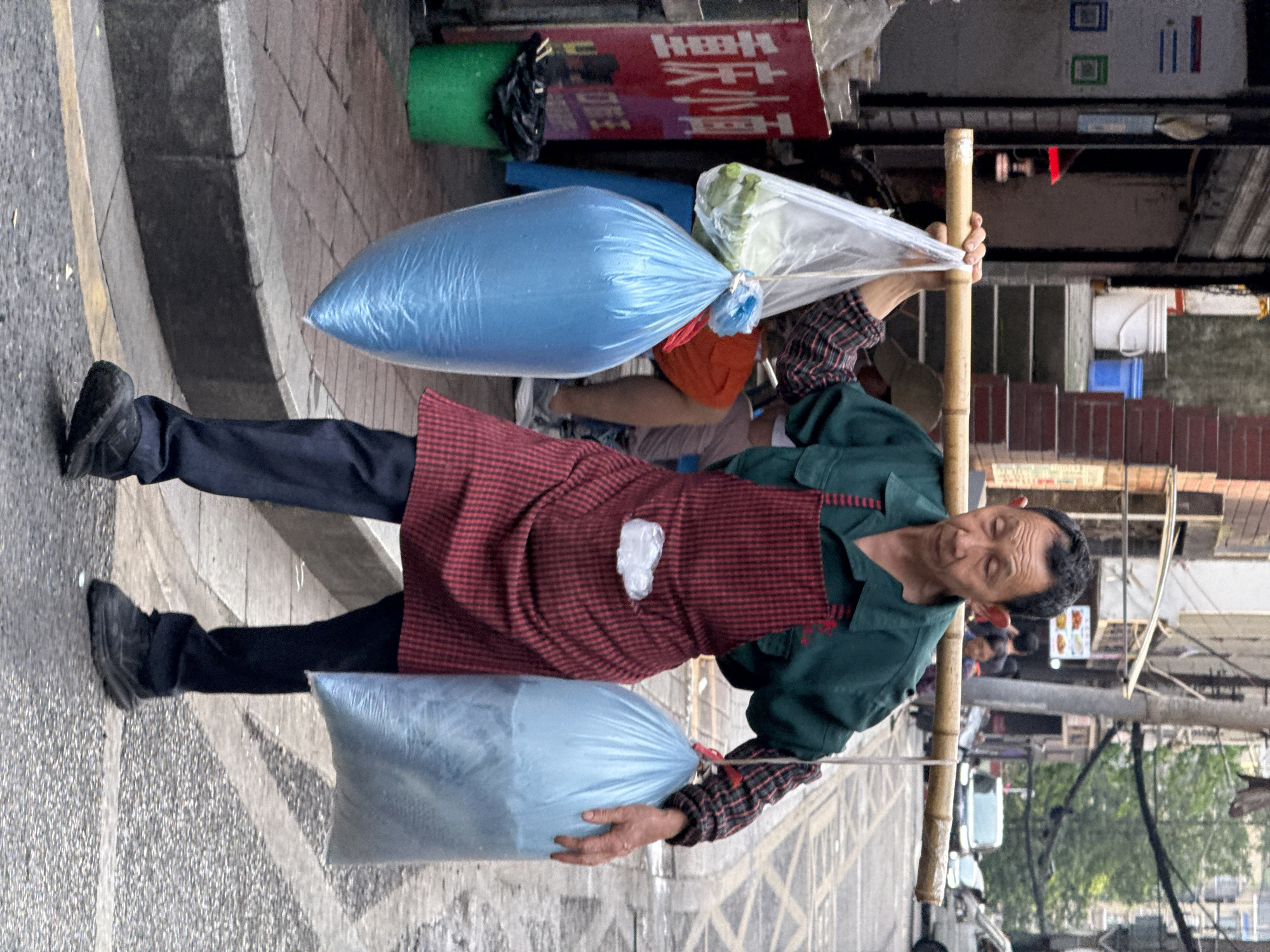 A middle-aged Asian man carries two large bags of goods on a bamboo pole across his shoulders. He wears a dark green jacket, a red and black plaid apron, and black pants and shoes. One bag appears to contain vegetables. He walks on a paved street in Yuzhong District, China, based on visible Chinese signage and architectural cues. People and cars are visible in the background, suggesting a busy urban area.