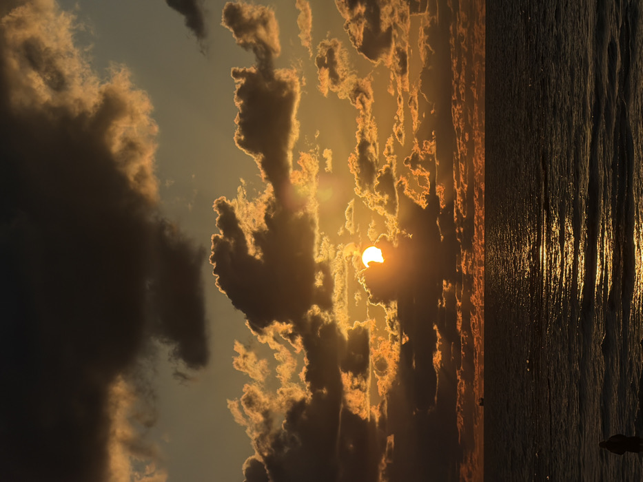 A vertical landscape captures a sunset over water. Dense, dark clouds occupy the left, contrasting with lighter, textured clouds on the right where the sun, partially obscured, emits a bright glow. Light rays pierce through the clouds, illuminating the horizon. The water reflects the sunlight, creating a shimmering, vertical band. A distant figure stands silhouetted near the water's edge.