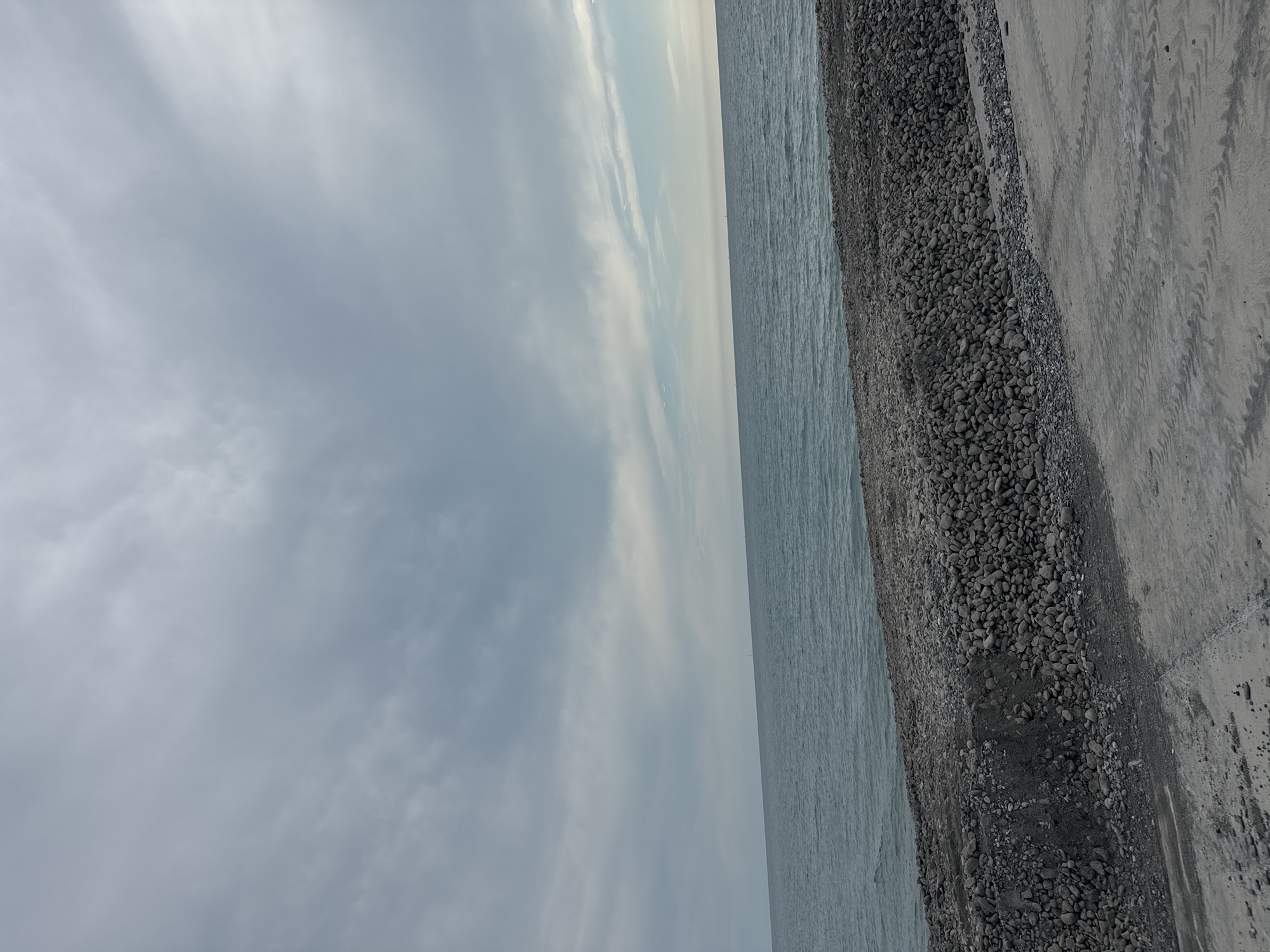 The image depicts a serene, overcast day at the seashore in Sirius, Russia. The horizon is a clean line separating the calm, grey-blue water of the Black Sea from a soft, cloudy sky. In the foreground, a rocky shoreline transitions to a sandy area with tire tracks from a vehicle, suggesting recent activity. The overall atmosphere is one of quiet contemplation, with no people or prominent man-made structures visible, save for the faint outlines of distant sailboats on the water.