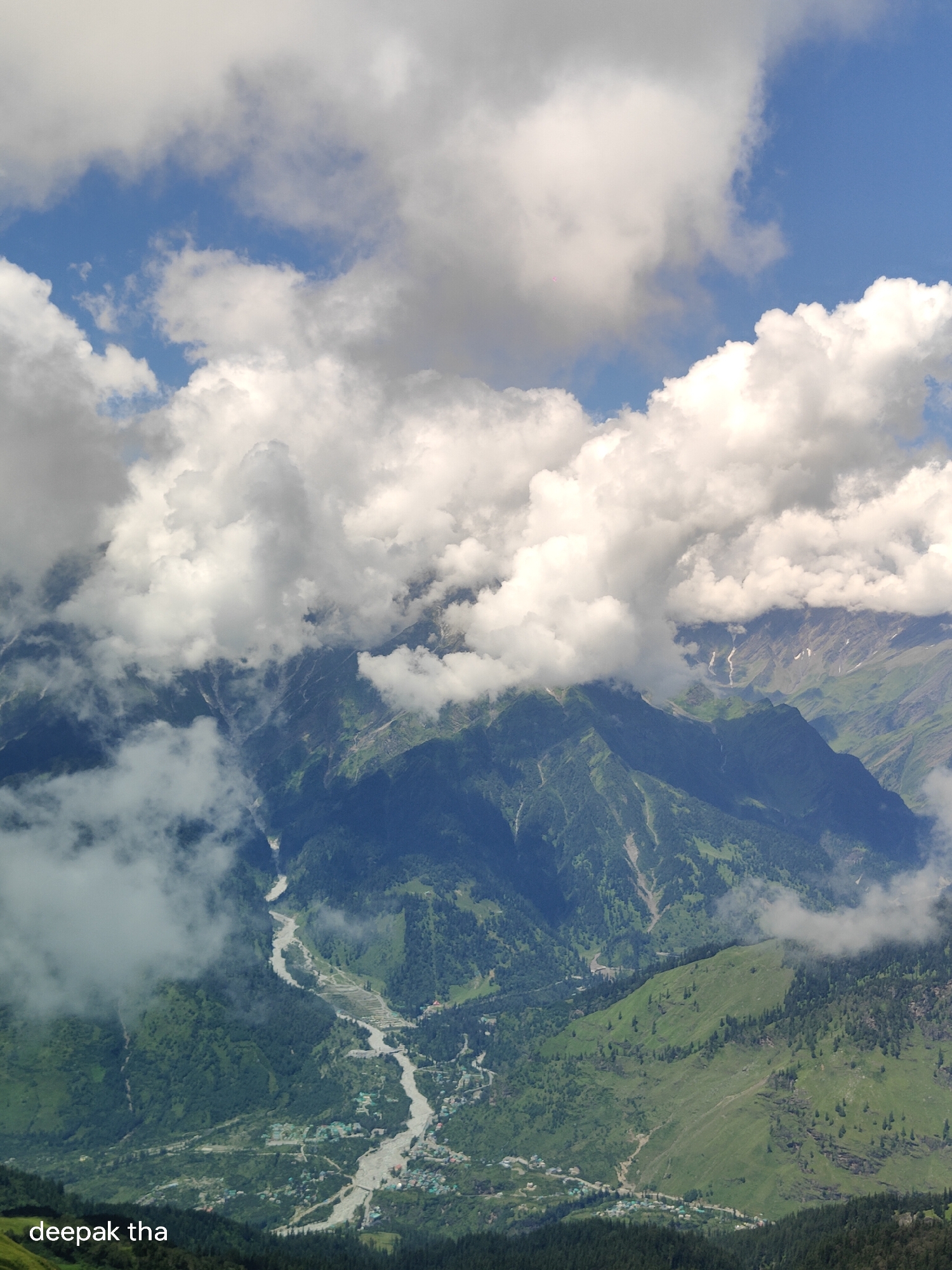 This is an aerial, vertical photograph of a mountainous landscape in the city of Bashisht, India. The sky is a vibrant blue, with large, fluffy white clouds dominating the upper portion of the image. These clouds partially obscure the peaks of the mountains, creating a dramatic, layered effect.
The mountains themselves are covered in lush green vegetation, with patches of darker green suggesting dense forests. A winding river, appearing light brown or grey, cuts through the valley floor, reflecting the overcast sky. Along the river, small clusters of buildings, indicative of a settlement, are visible. The terrain is steep and rugged, with visible rock formations and scree slopes in some areas. The image captures a sense of natural grandeur and the remote beauty of the Indian Himalayas. A watermark at the bottom left corner reads "deepak tha".