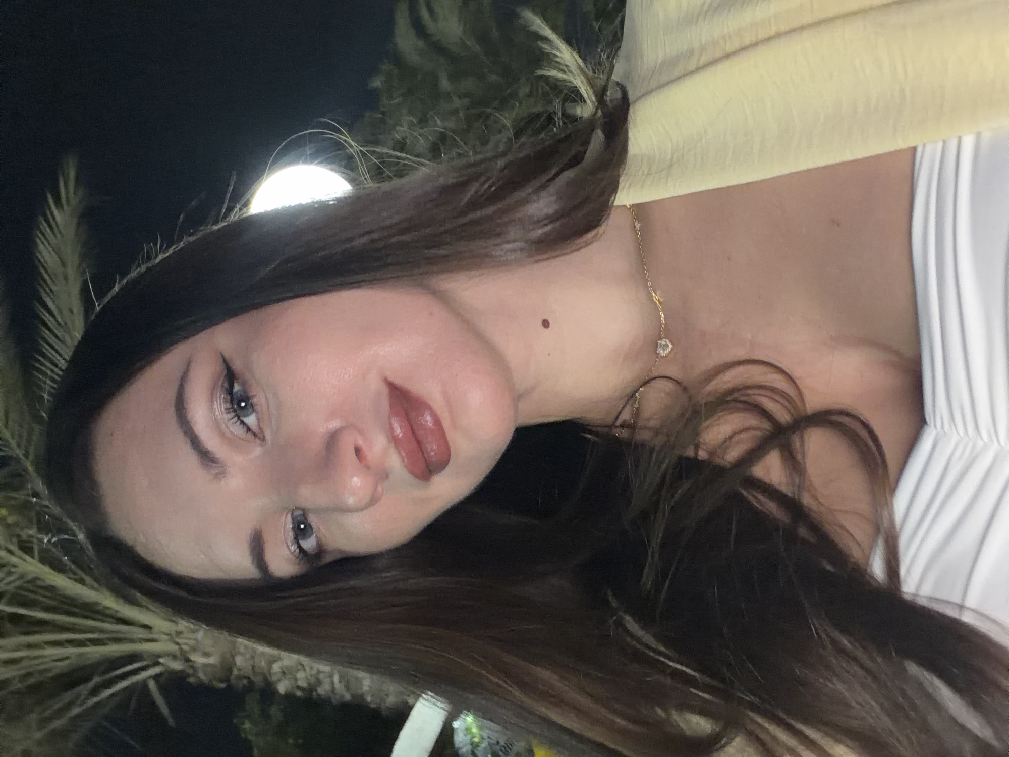 The image is a close-up, outdoor, night-time selfie of a woman with long, dark brown hair. She has fair skin, blue eyes with dark eyeliner, and her lips are painted with a dark, glossy lipstick. She is wearing a strapless white top and a light yellow jacket or shawl. A delicate gold necklace with a heart-shaped pendant adorns her neck. A small mole is visible on her neck below her jawline. The background is dark, with hints of palm trees and artificial lighting, suggesting an urban or resort setting. The overall impression is of a posed portrait taken at night, possibly in a warm climate. The location of 6th of October, Egypt is not explicitly visible in the image itself but may be inferred from external information not provided with the image.