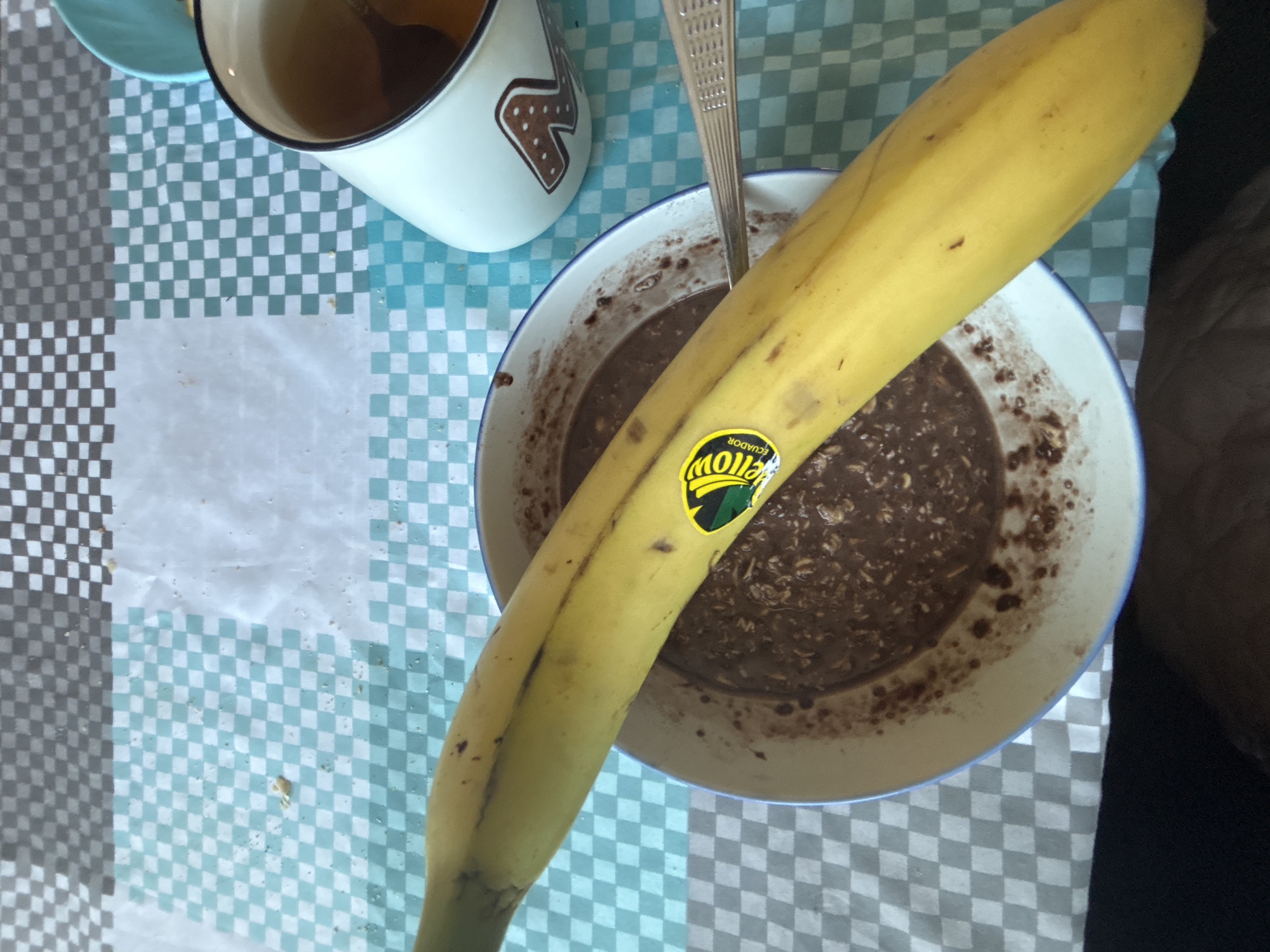 The image shows a breakfast scene. On a checkered tablecloth, there's a mug with a brown liquid, likely tea or coffee, and a bowl filled with a dark, porridge-like substance, possibly oatmeal. A banana with a Chiquita sticker lies across the bowl. A spoon rests inside the bowl. There are no people visible in the image. The location in the city of Zlatograd, Bulgaria is not discernable from this image alone.