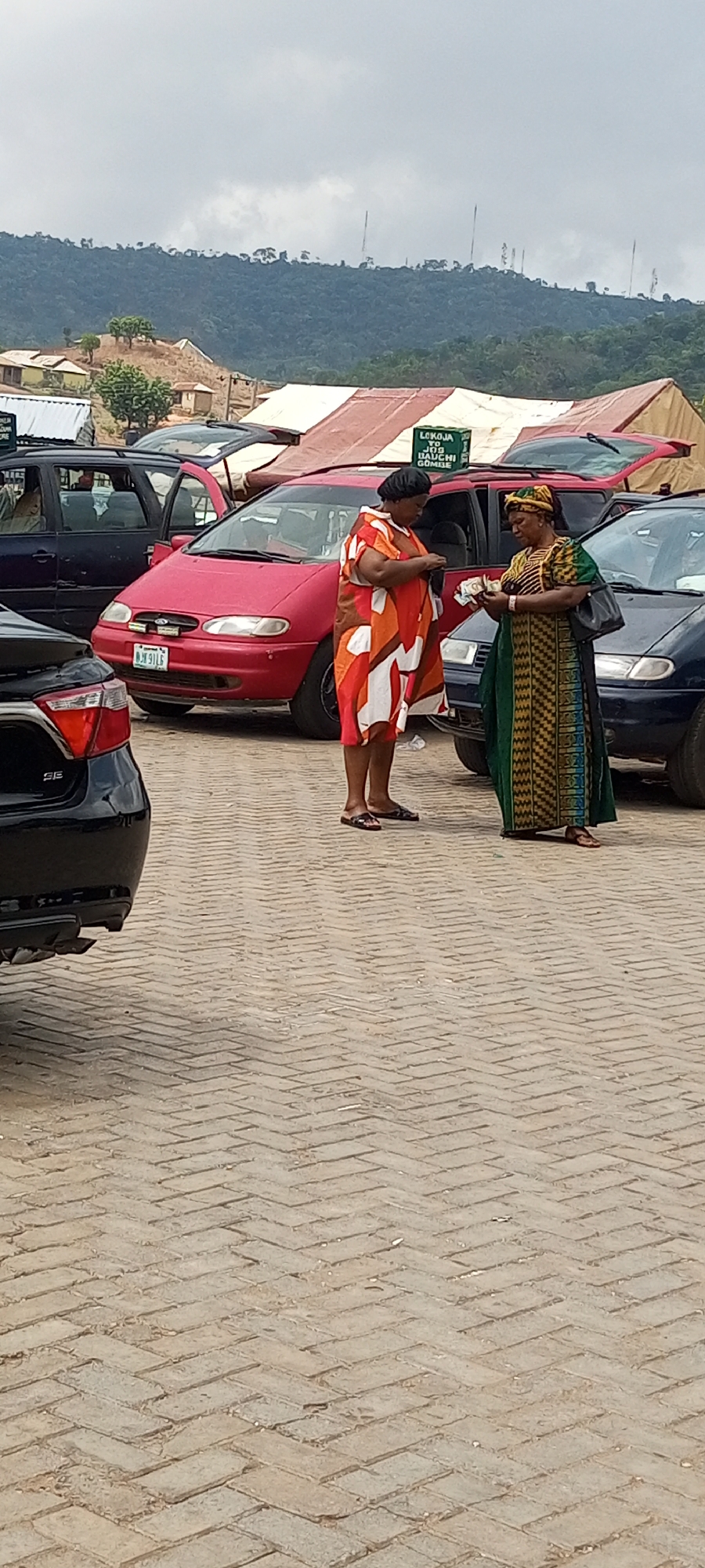 Two African women stand close together on a paved area amidst parked cars in Lokoja, Nigeria. The woman on the left wears a multicolored dress with orange, red, and white patterns and dark sandals. She is holding and looking at money in her hands. The woman on the right is wearing a long green and gold patterned dress with a yellow headwrap, and she also holds currency in her hands. In the background, there are several parked cars of various colors, including red, black, and blue. A sign is visible, partly obscured, which reads "Lokoja to..." The setting appears to be an outdoor parking area with a mountainous landscape in the distance under a cloudy sky. There are buildings on a hillside visible in the background.