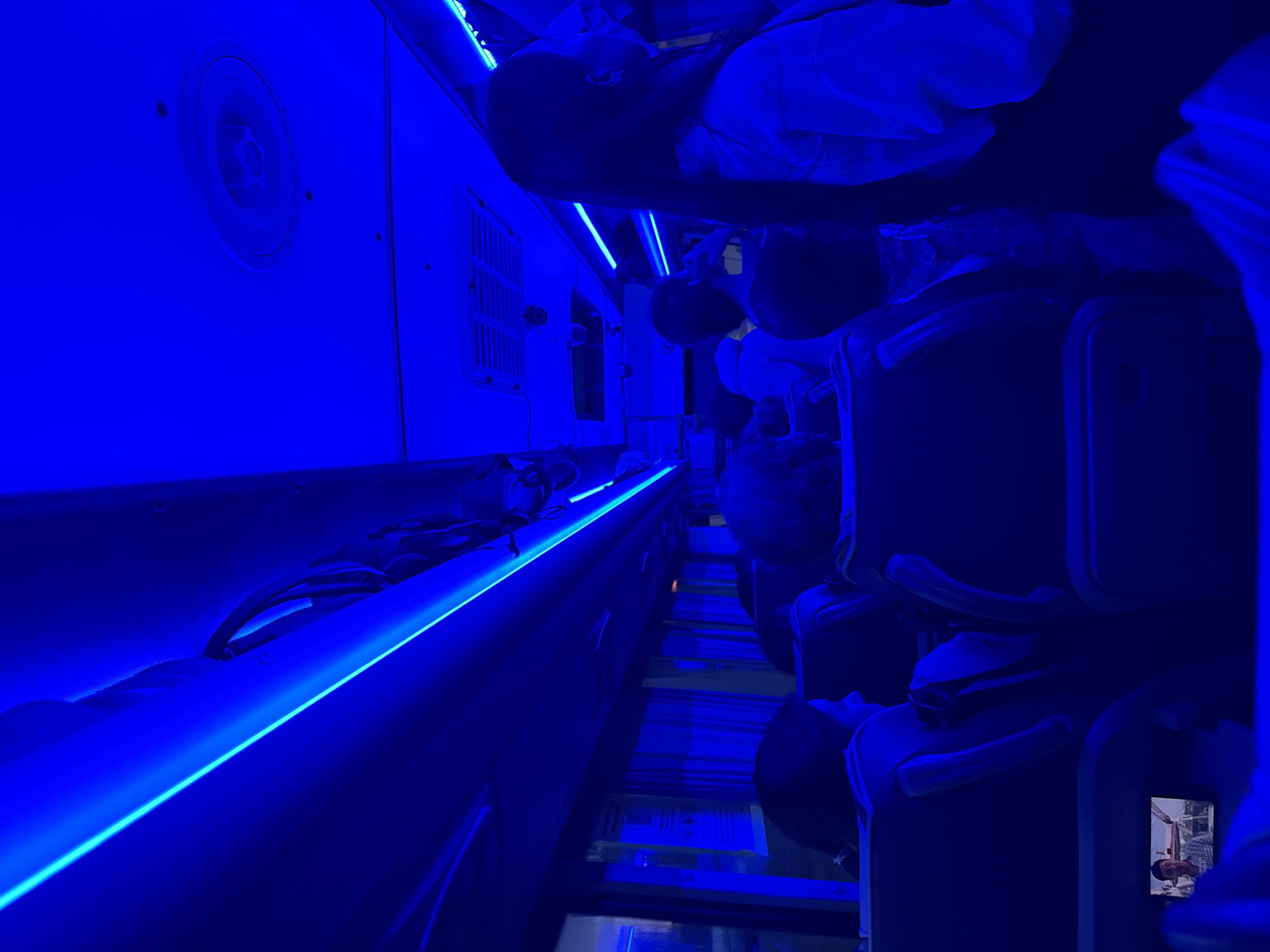 The image shows the interior of a bus at night, bathed in bright blue light from neon strip lights running along the ceiling. Several passengers are visible, seated in rows, facing forward. Some passengers are looking out the windows, while others appear to be talking or engaged with their phones. The windows are covered by light-colored curtains. The overall atmosphere is dim, with the blue light casting long shadows. The small screen of a device held by a passenger in the foreground shows a man in a checkered shirt. The scene is devoid of any specific architectural details that would definitively place it in Kazan, Russia, but the lighting suggests a modern public transportation vehicle.