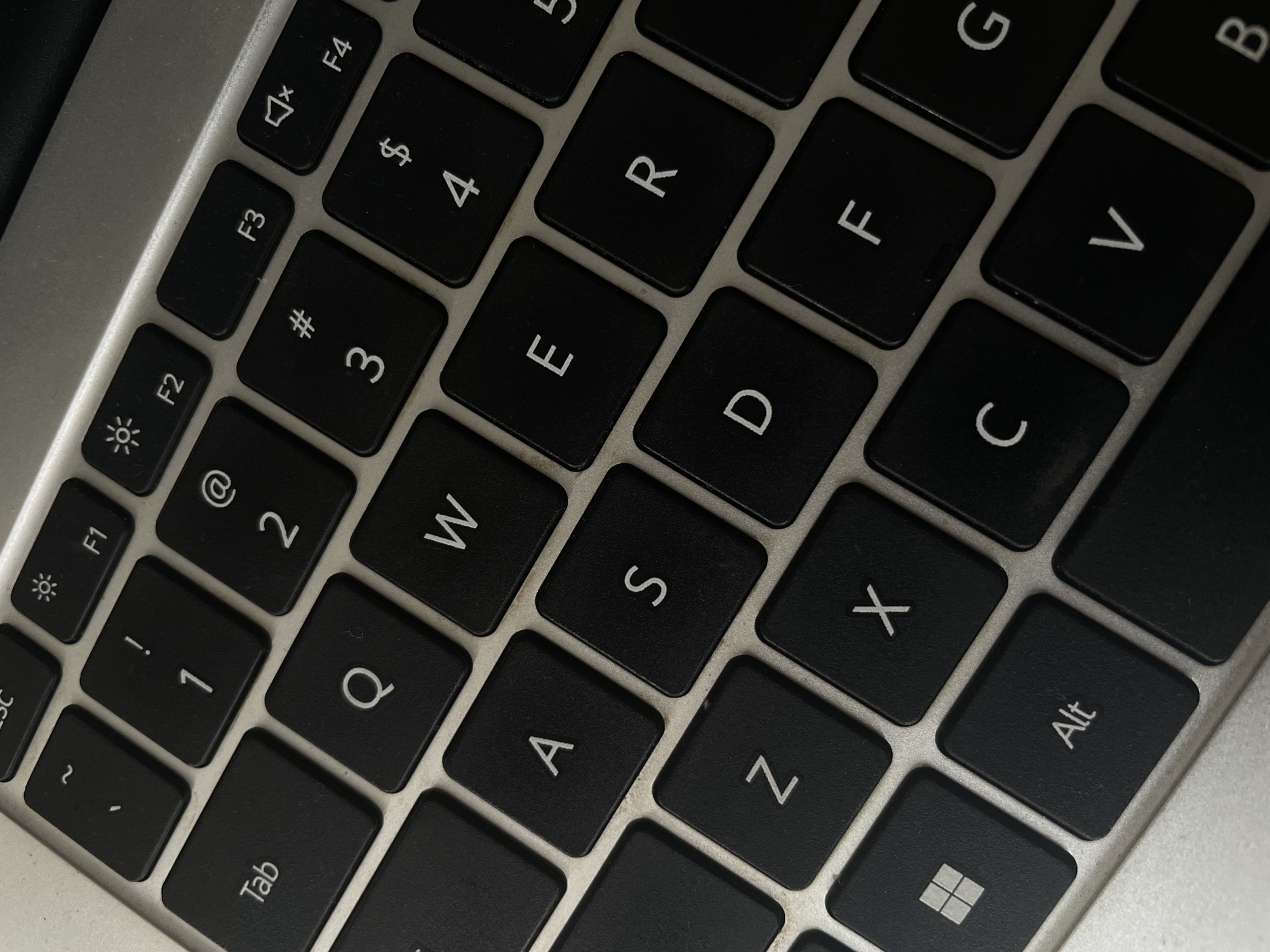 The image shows a close-up, high-angle view of a portion of a laptop keyboard. The keyboard is black with white lettering on the keys. Several keys are clearly visible, including "Tab", "Q", "A", "Z", "W", "S", "X", "E", "D", "C", "R", "F", "V", "G", "B", and several function keys ("F1" through "F4"). The lighting is somewhat dim, casting subtle shadows. There are no people or any identifiable location elements, such as a city, present in the image.