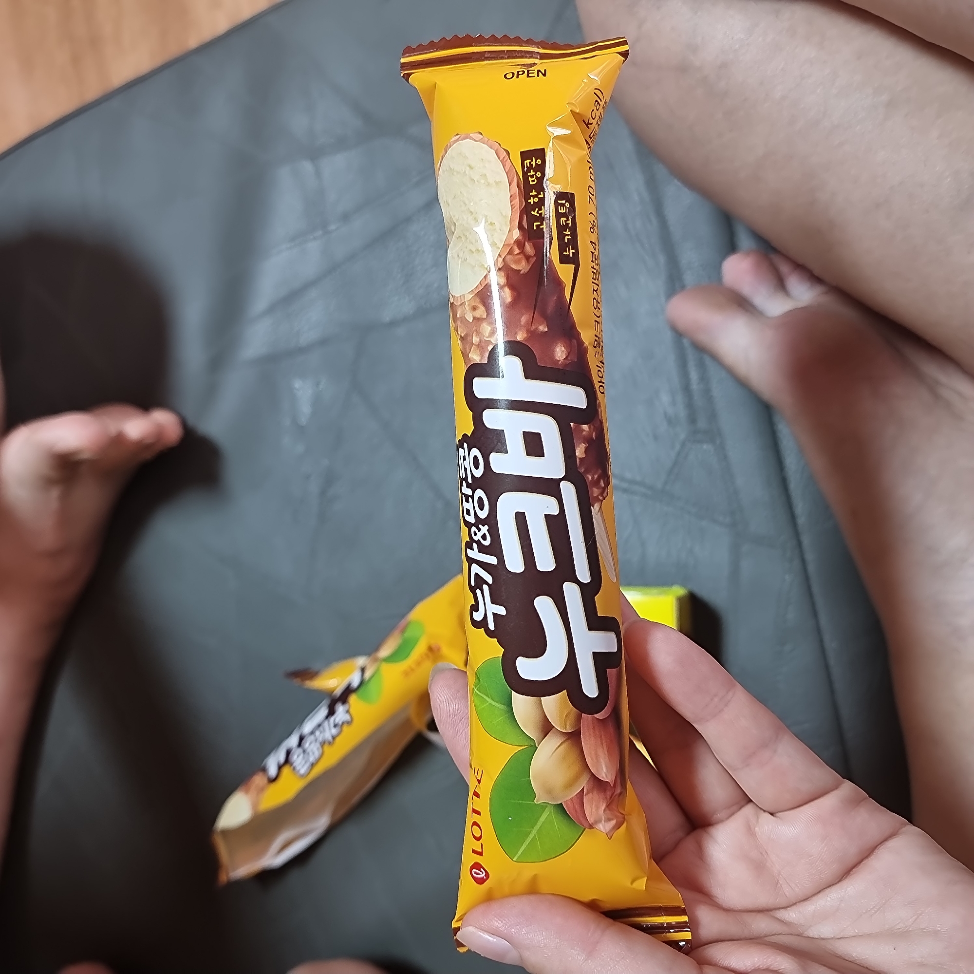 A person is holding a yellow chocolate bar with the brand name "LOTTE" visible. The chocolate bar has an image of peanuts and leaves. The text on the bar is in Korean and appears to say "누 가 & 땅콩" which translates to "Nougat & Peanuts" and "누크바" which means "Nuke Bar". The bar is partially unwrapped, revealing the chocolate coating with nuts. In the background, another partially unwrapped chocolate bar is visible, as are parts of a person's feet and legs, suggesting they are sitting or lying down. The environment appears to be indoors, on a grey surface. The image does not contain information about the city of Dunnae-myeon, South Korea.