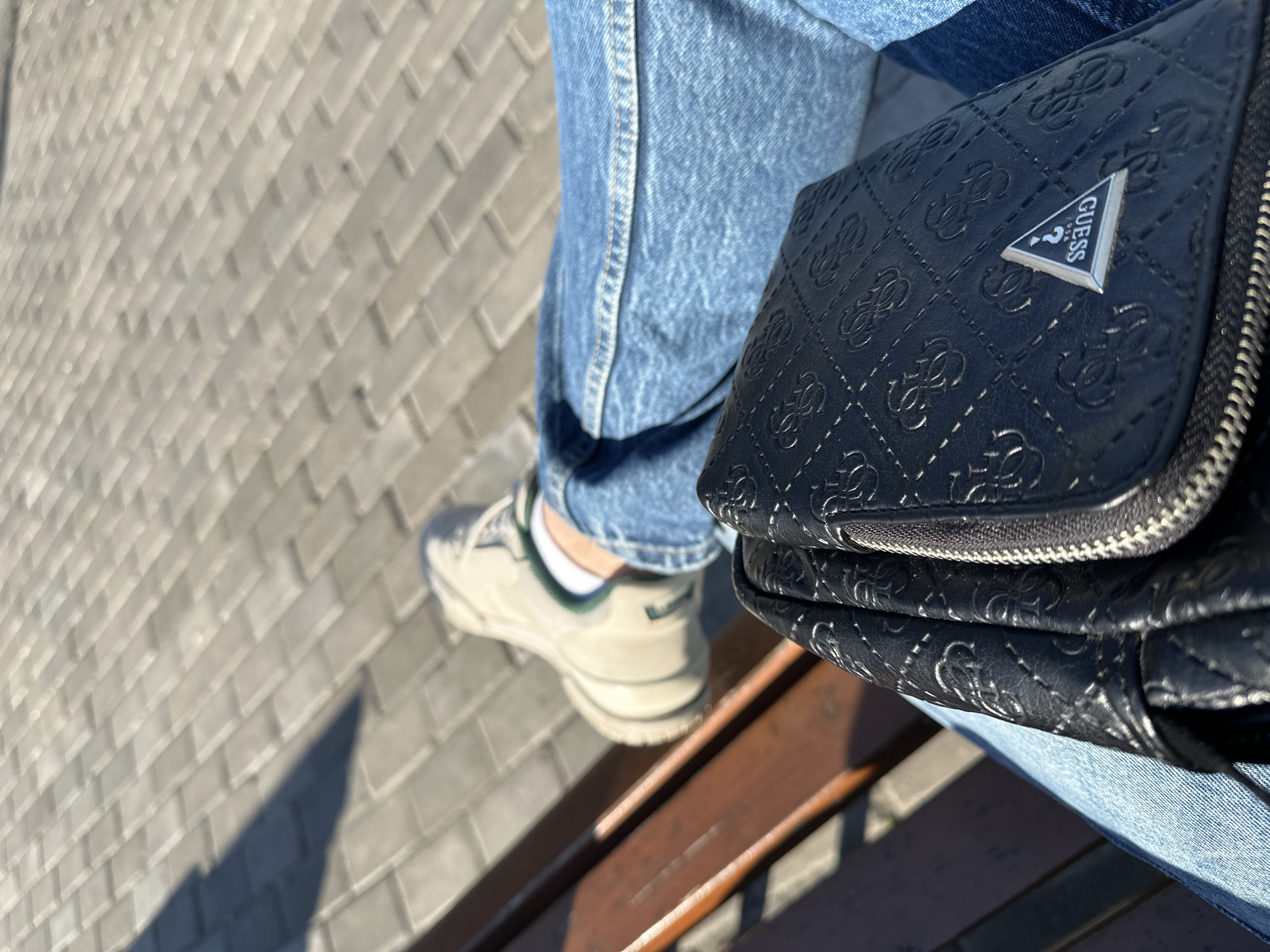 The image captures a close-up shot of a person seated on a bench, their leg crossed, with a black Guess purse resting on their lap. They are wearing light blue jeans and off-white sneakers with green accents. The bench appears to be made of brown wood. The background consists of gray interlocking pavers, typical of a city sidewalk or plaza. The time of day seems to be sunny, as indicated by the shadows on the pavers. The location of the scene is Unknown, Unknown.