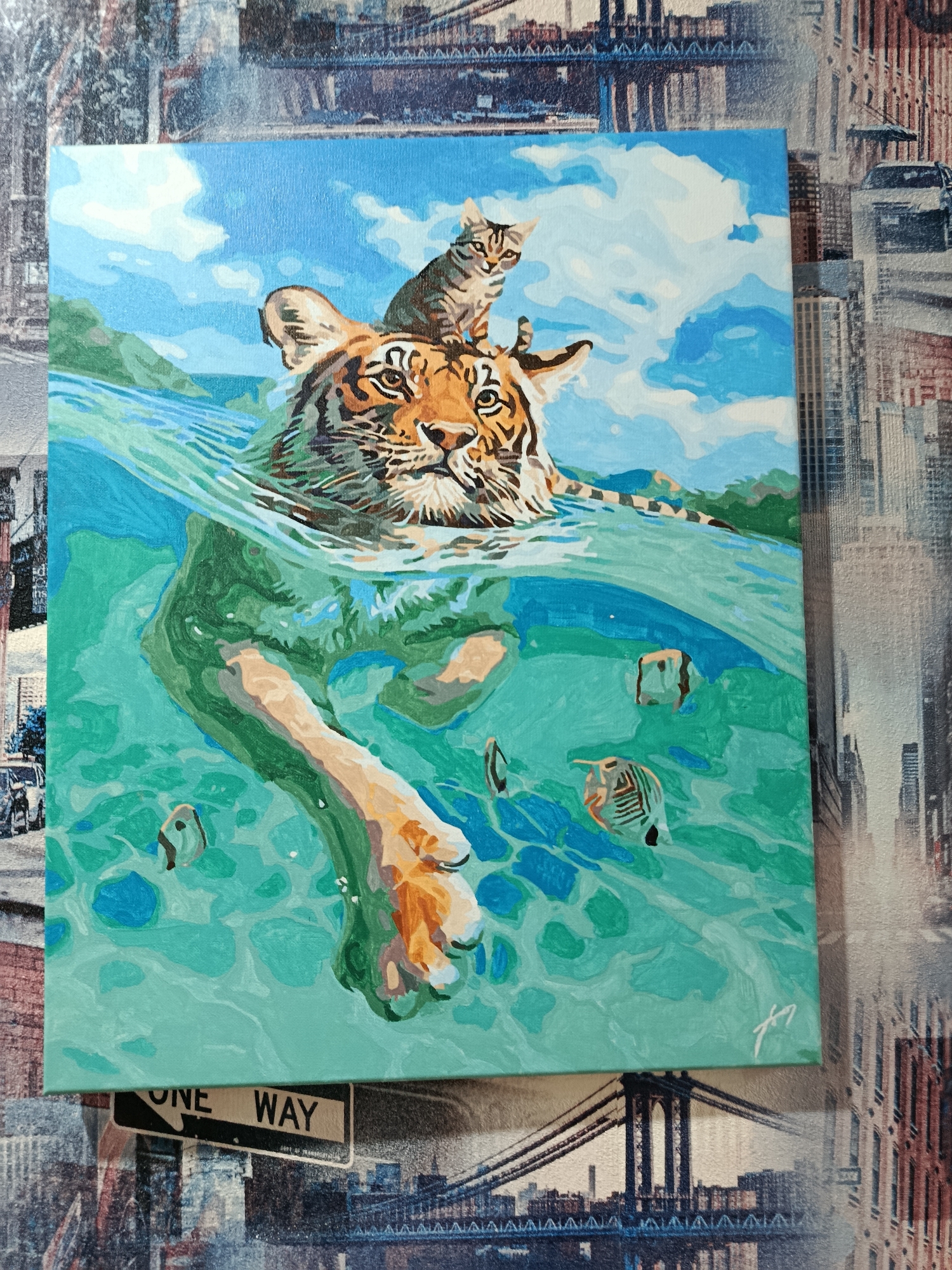 The image shows a painting of a tiger swimming in turquoise water with a cat sitting on its head. The tiger's head and front paws are visible above the water, while its body and hind legs are submerged. There are several small fish swimming around the tiger. The background depicts a landscape with green hills and a blue sky with white clouds. A signature appears in the lower right corner of the painting. The painting is placed against a backdrop featuring a collage of cityscapes, including bridges and buildings, and a "ONE WAY" street sign. There is no visible connection to the city of Apatity, Russia, in this image.