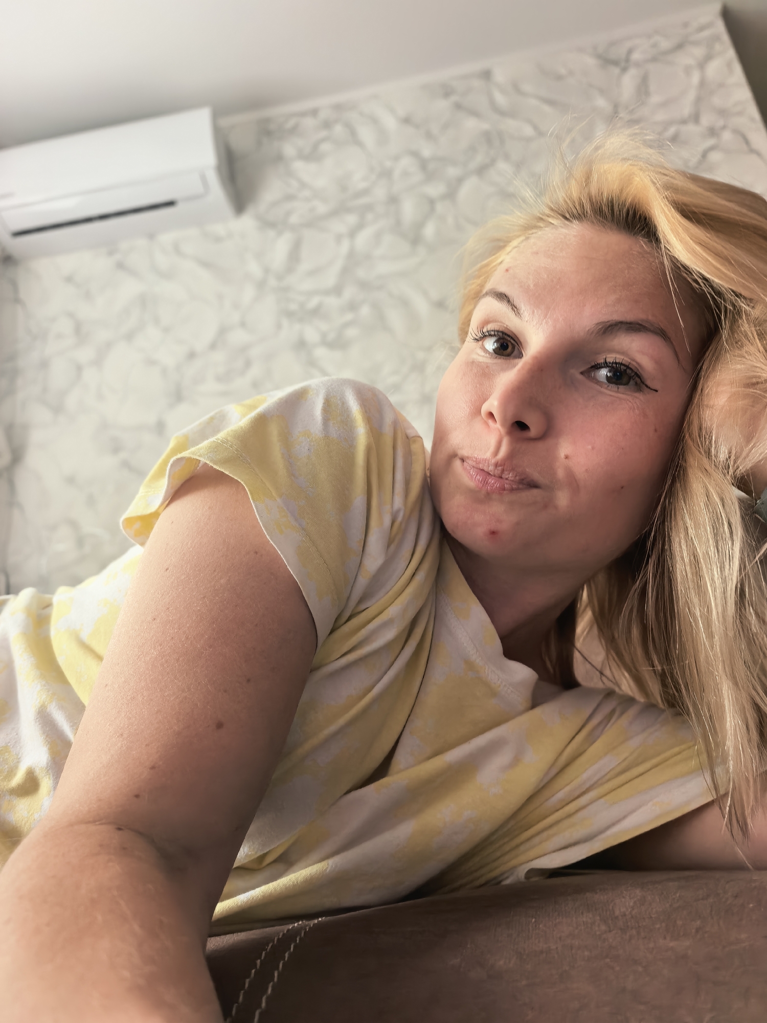 The image shows a young woman taking a selfie indoors, presumably in a residential setting in Sterlitamak, Russia. She has blonde hair, light skin, and light brown eyes, looking directly at the camera with a soft, slightly pursed-lip expression that conveys a contemplative or mildly amused mood. Her head is tilted, and her left arm, covered by a yellow and white patterned or tie-dye t-shirt, appears to be supporting her.
In the background, a white split-system air conditioning unit is mounted on a wall covered with a light, subtly marbled or textured wallpaper. The lighting is bright and even, suggesting it is daytime. Notable details include a few small red blemishes on her chin and forehead, and light freckles across her nose and cheeks. She has light eyeliner applied to her upper eyelids. Her blonde hair is casually disheveled, falling around her face and over her shoulders. A brown, possibly upholstered, surface occupies the lower foreground, suggesting she might be lying down or leaning back on a couch or bed. There is no visible text within the image. The scene conveys a relaxed and casual moment.