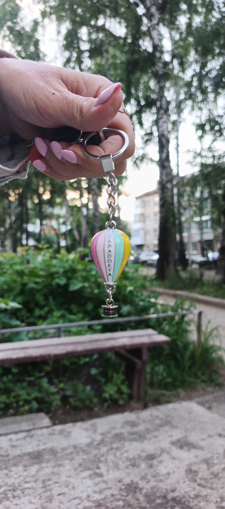 A close-up shot focuses on a person's hand holding a keychain with a hot air balloon charm. The background depicts a blurred outdoor scene with trees, a park bench, and residential buildings, suggesting an urban or park setting, possibly in the city of Chaikovsky, Russia. The person's nails are painted light pink, and they are wearing a ring on their right hand. The hot air balloon charm is colorful, with segments in pink, yellow, and mint green, and appears to have the word "CAPADOKYA" written vertically on one of its sides. Below the balloon is a small, dark-colored basket. The lighting suggests it is daytime, with a soft, diffused light.