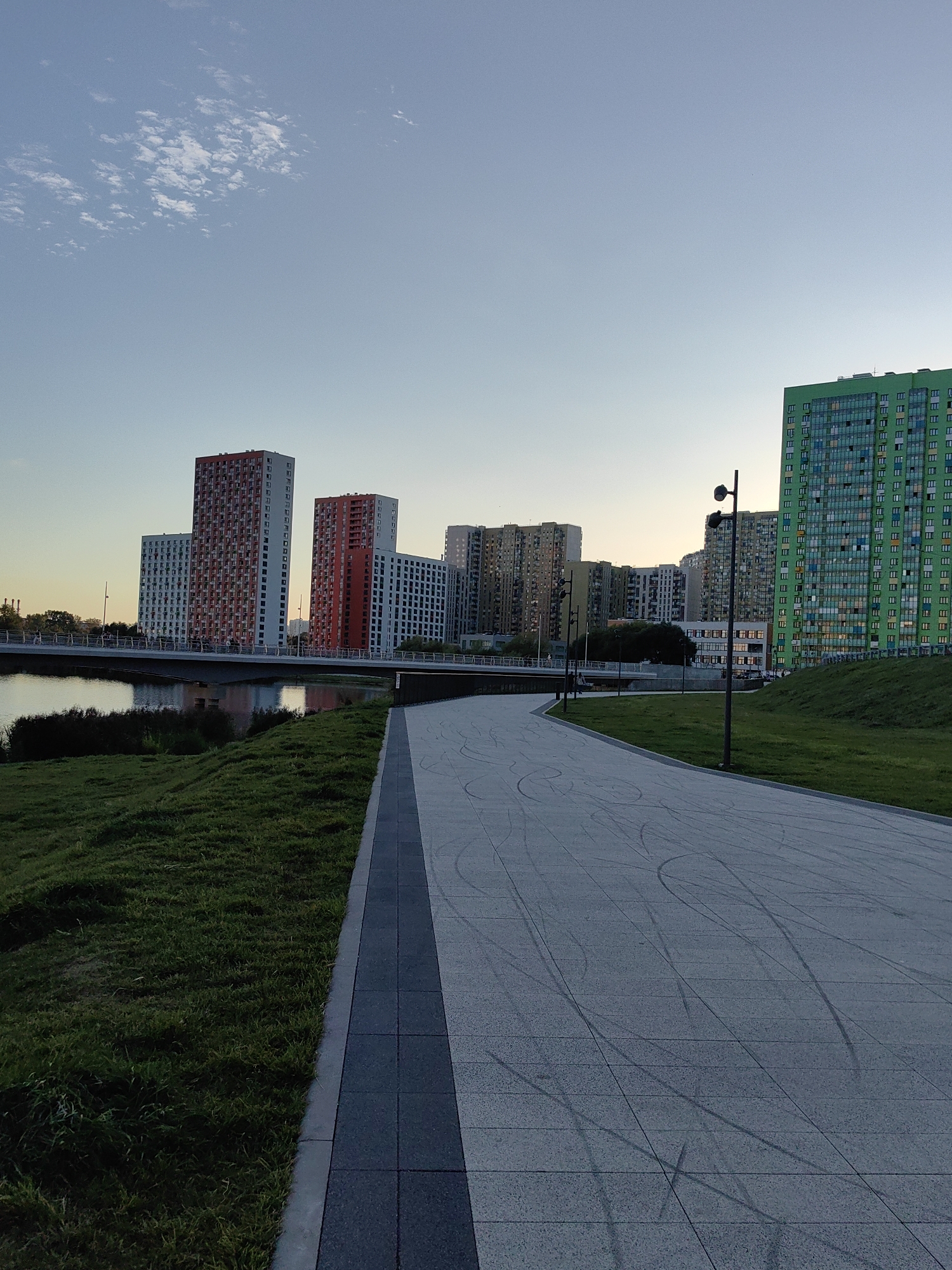 This is a wide shot of a modern cityscape with a river in the foreground. The sky is a pale blue with a few wispy clouds. In the distance, several tall, multi-story residential buildings are visible, some with red and white facades, others in muted earth tones, and one striking green building. A bridge spans the river, connecting the two sides of the city. A paved walkway runs along the riverbank, lined with green grass. The walkway has tire marks on it, suggesting recent activity. Several lampposts are positioned along the walkway. The overall impression is of an urban park area alongside a residential development. This scene is located in Moscow, Russia, specifically within the Zamoskvorechye district.