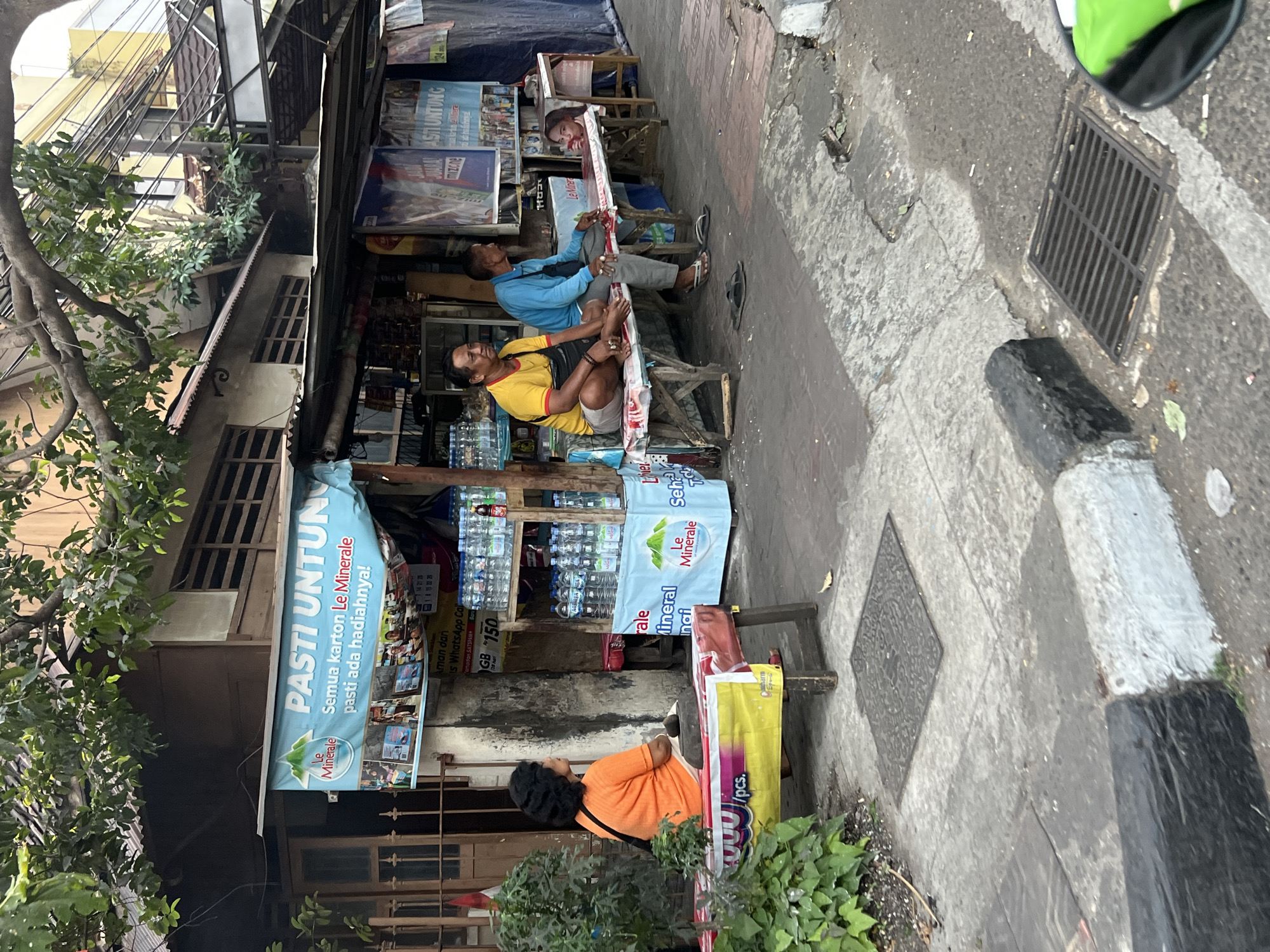 The image captures a street scene in Yogyakarta, Indonesia, featuring a small roadside stall selling bottled water. In the foreground, a woman with dark hair, wearing an orange t-shirt, sits on a bench. To her right, two men are seated on another bench, one in a yellow shirt and the other in a blue shirt. A young girl is partially visible behind the man in the blue shirt. The stall displays an assortment of bottled water, with prominent signage in Indonesian for "Le Minerale". The setting appears to be an urban environment, with a sidewalk, street, and buildings in the background. The time of day is not explicitly indicated, but the lighting suggests it could be daytime.