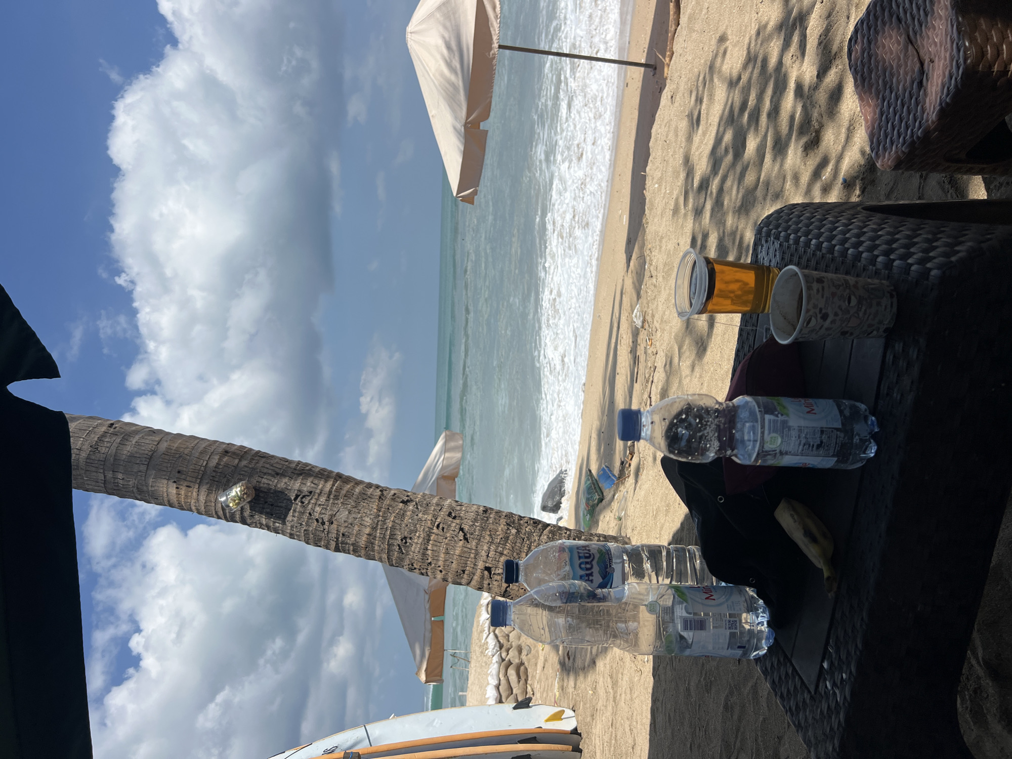 This image depicts a beach scene in Kuta, Indonesia, characterized by a sandy shore and the ocean under a bright, cloudy sky. In the foreground, a dark-colored woven table holds several items: two plastic bottles of Aqua brand bottled water, a black hat, a peeled banana, and two cups, one containing a light brown liquid (likely beer or iced tea) and another with a dark liquid (likely coffee). To the right of the table, a small brown woven stool is visible.
Behind the table, a large palm tree trunk stands prominently. Attached to the trunk at eye level is a small, clear plastic container filled with what appears to be snacks or nuts. Further back on the sand, several objects are scattered: a large white surfboard on the left, two sandbag barriers, a beach umbrella, and another beach umbrella further in the distance. The ocean waves are breaking on the shore, creating a foamy white line. The overall atmosphere suggests a relaxed, tropical setting.