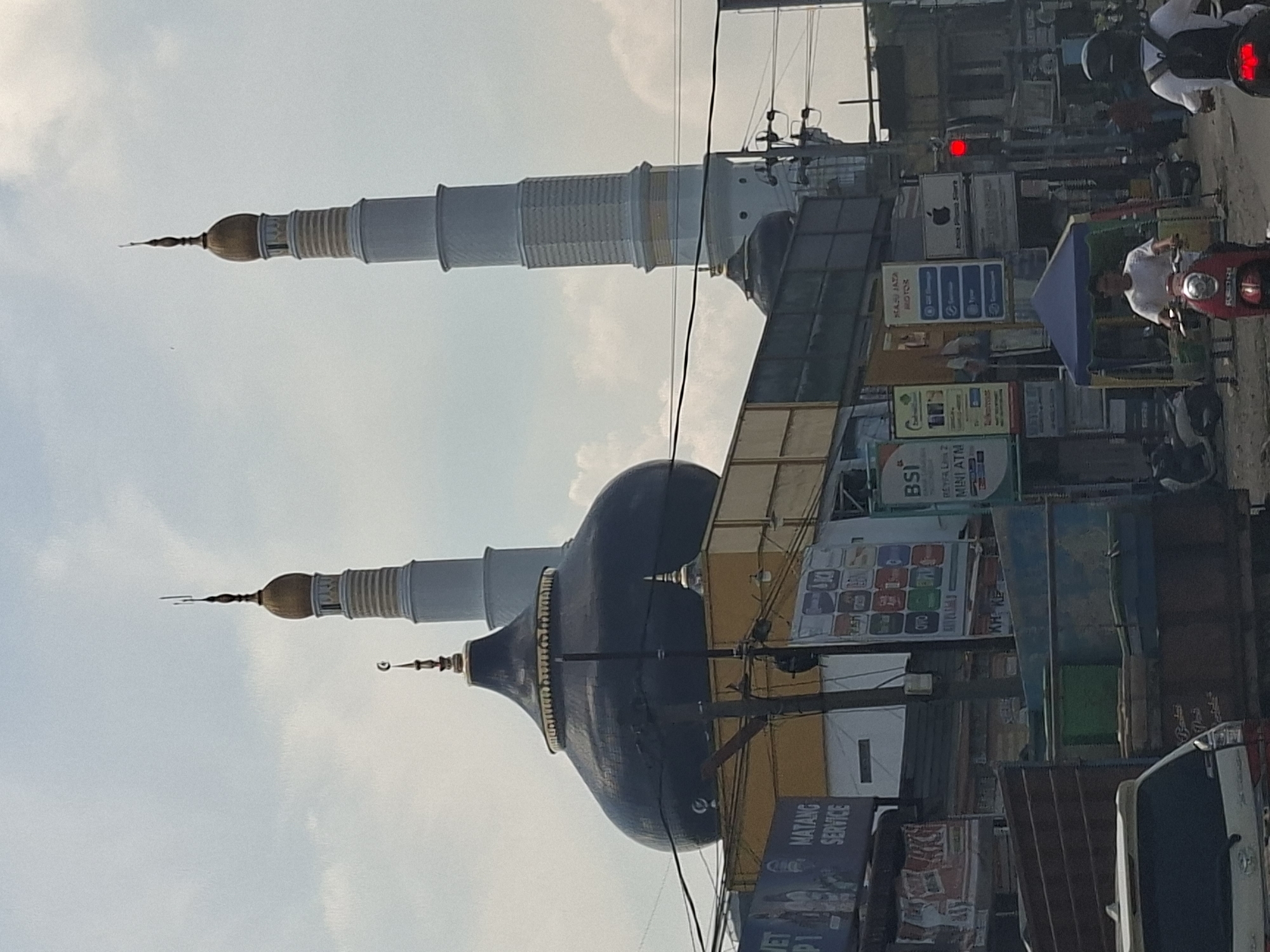 The image shows a street scene in Pantegajah, Indonesia, with a mosque prominently featured. The mosque has a large, dark dome and two tall minarets. The left minaret is white with gold accents, and the right minaret is also white with gold bands. In the foreground, there are several vehicles, including a truck, a car, and a motorcycle with a rider. A person on a red scooter is visible in the center of the frame, wearing a white shirt. To the right, another person on a motorcycle, wearing a white shirt and black backpack, is partially visible. Various signs and advertisements are displayed on buildings along the street, including one for "MATANG SERVICE" and another for "BSi MINI ATM". The sky is overcast with some clouds. The scene depicts a busy urban environment.