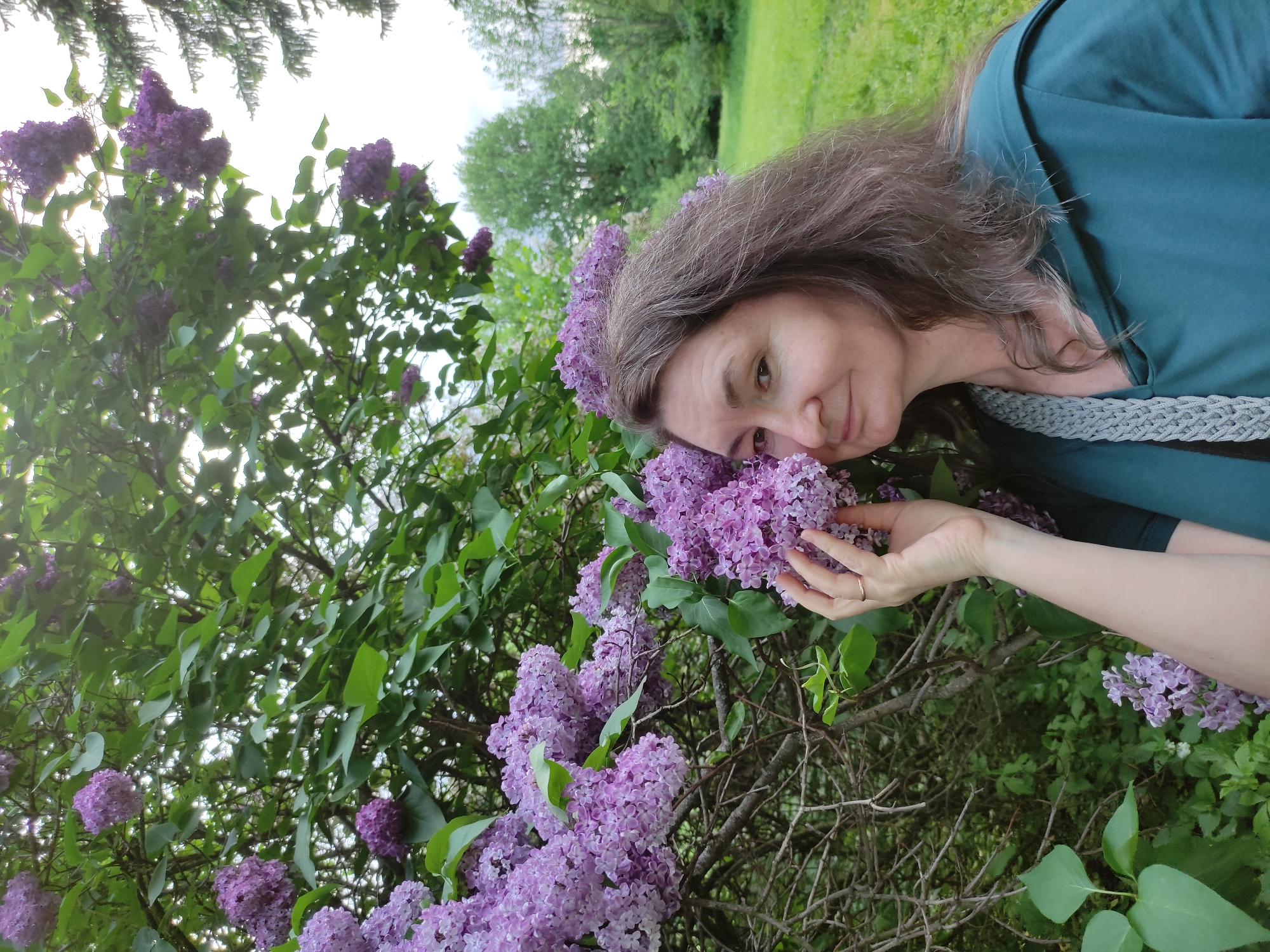 A middle-aged woman with long brown hair and a green shirt is seen close-up, her face near a blooming lilac bush. She is holding the flowers. The background includes green trees and a hint of sky. The exact location within Moscow, Russia is not identifiable from the visual information provided.