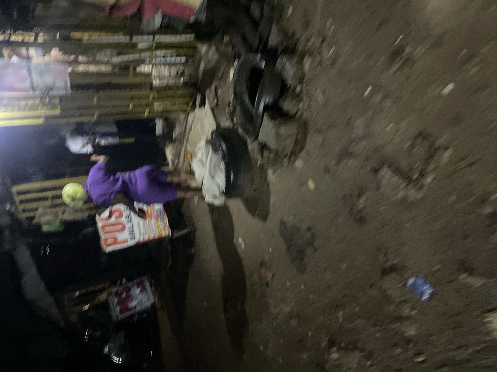 The image depicts an outdoor scene at night, likely in a commercial area of Abuja, Nigeria. In the foreground, the ground is uneven and appears to be dirt or mud, with scattered debris including a blue wrapper. Two old tires are visible, one lying flat and the other propped up on blocks, suggesting a makeshift seating area or storage.
In the middle ground, a person wearing a purple outfit and a yellow head covering is walking away from the camera. Beside them, a sign with "POS" prominently displayed indicates a point-of-sale service. Further back, a dark structure resembling a stall or shop is visible, with a yellow metal gate. Silhouettes of other individuals can be seen inside or behind the stall, suggesting ongoing activity.
The overall lighting is dim, with artificial light sources from the stall illuminating the area. The environment appears somewhat unkempt, with a mix of commercial signage, discarded items, and rudimentary infrastructure.