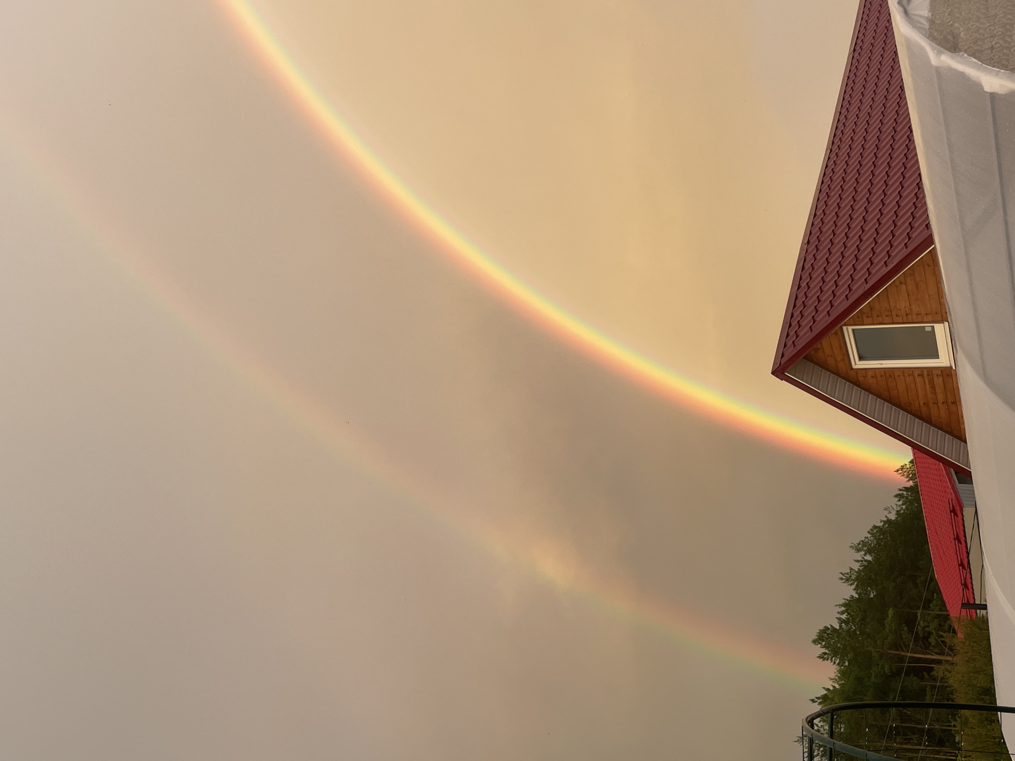 The image captures a double rainbow arcing across a hazy, pale orange sky above a rural scene. In the foreground, the dark red, corrugated metal roof of a house with wooden siding and a single window is visible. A white, translucent structure, possibly a greenhouse, is partially visible in the lower right corner. Behind the house, a dense line of dark green coniferous trees forms the horizon. The sky is dominated by the vibrant colors of two superimposed rainbows, with the lower, brighter rainbow displaying the full spectrum of colors from red to violet, and a fainter, secondary rainbow appearing above it. The overall impression is one of a natural phenomenon occurring in a quiet, possibly remote, residential area. The location is identified as Zav'yalovo, Russia.