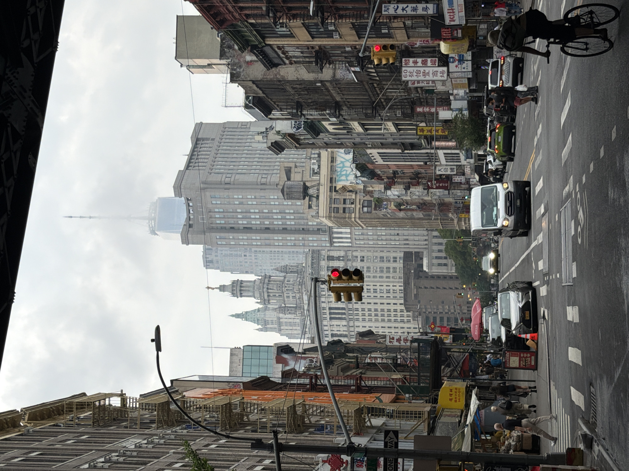 A wide shot captures a street scene in New York City, likely in Chinatown, under an overcast sky. The foreground shows a pedestrian crossing with a cyclist in the right foreground. Traffic lights are visible, with one displaying a red light. Cars and a white van are on the road. Buildings line both sides of the street, featuring fire escapes and various signs, some in Asian script, suggesting the location. In the background, skyscrapers of varying heights dominate the skyline, with a prominent spire-like structure and another taller building possibly being the One World Trade Center. The overall atmosphere is urban and bustling, despite the subdued lighting.