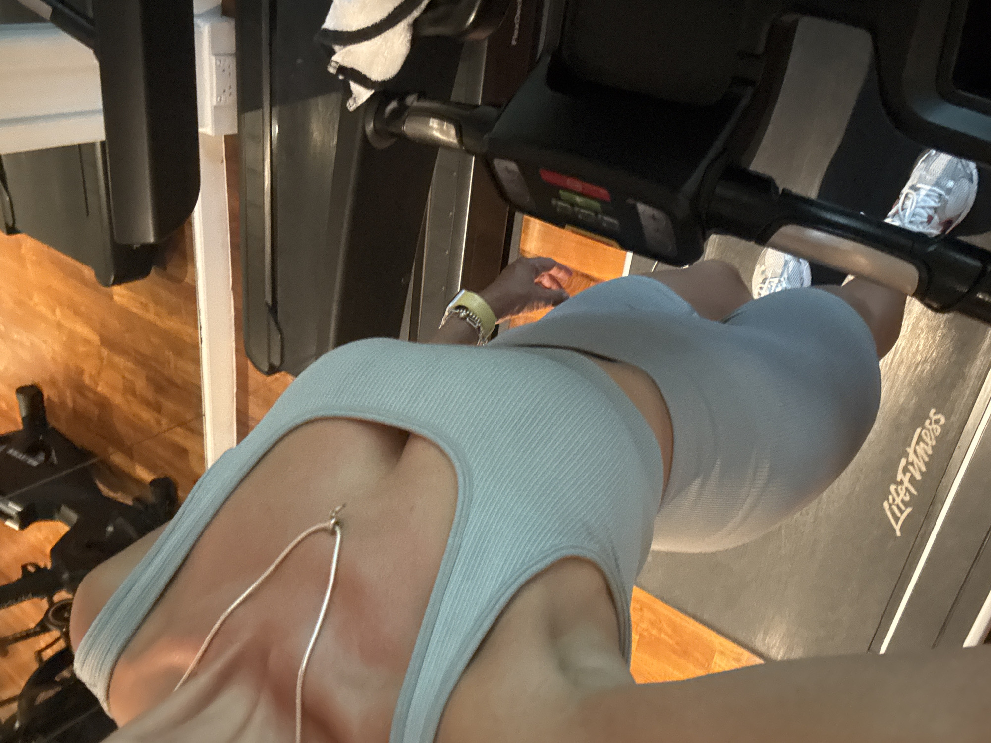 This high-angle, top-down perspective image captures a person standing on a treadmill, likely in a home gym or fitness area in Kuta Selatan, Indonesia. The person is wearing a light blue or grey ribbed sports bra/crop top with a subtle cut-out design, paired with matching ribbed shorts. A slender silver chain necklace drapes down their chest.
On the person's left wrist, a bright yellow wristband or watch is visible, accompanied by several silver bracelets. Their hands are placed on the black handlebars of the treadmill. The person is wearing white athletic shoes with grey accents and white laces.
The treadmill itself features a black track and a control panel with red and green lights. The brand name "LifeFitness" is prominently displayed in white text on the treadmill belt, and "FlexDeck" is partially visible on the console. The surrounding area includes light brown wooden flooring and a white wall with an electrical socket. A white towel with black stripes is draped over a part of the treadmill's frame. The scene is brightly lit, indicating an indoor setting.