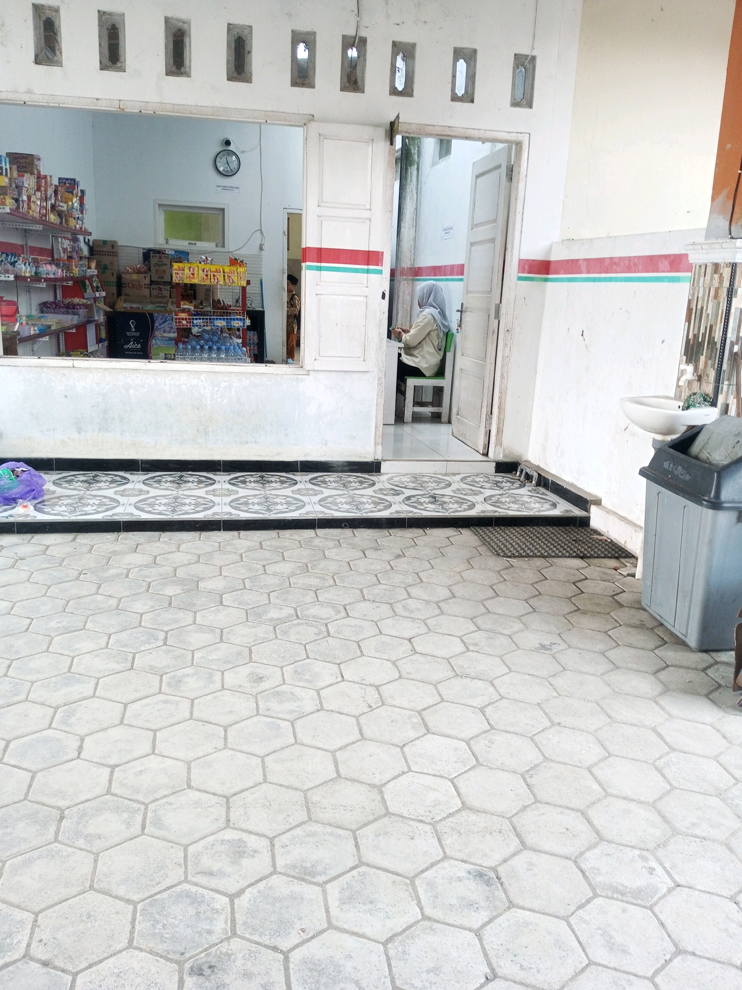 The image shows the exterior of a small shop in Kalibeber, Indonesia. A woman wearing a light-colored hijab and a tan jacket is seated at a table inside the shop, facing a doorway. Shelves stocked with various goods are visible through an open window in the shop. Outside the shop, there is a tiled floor area with hexagonal patterns. A grey trash bin and a sink are located near the doorway. The shop has white walls with decorative red and green stripes along the exterior. The floor outside is made of light gray hexagonal tiles.