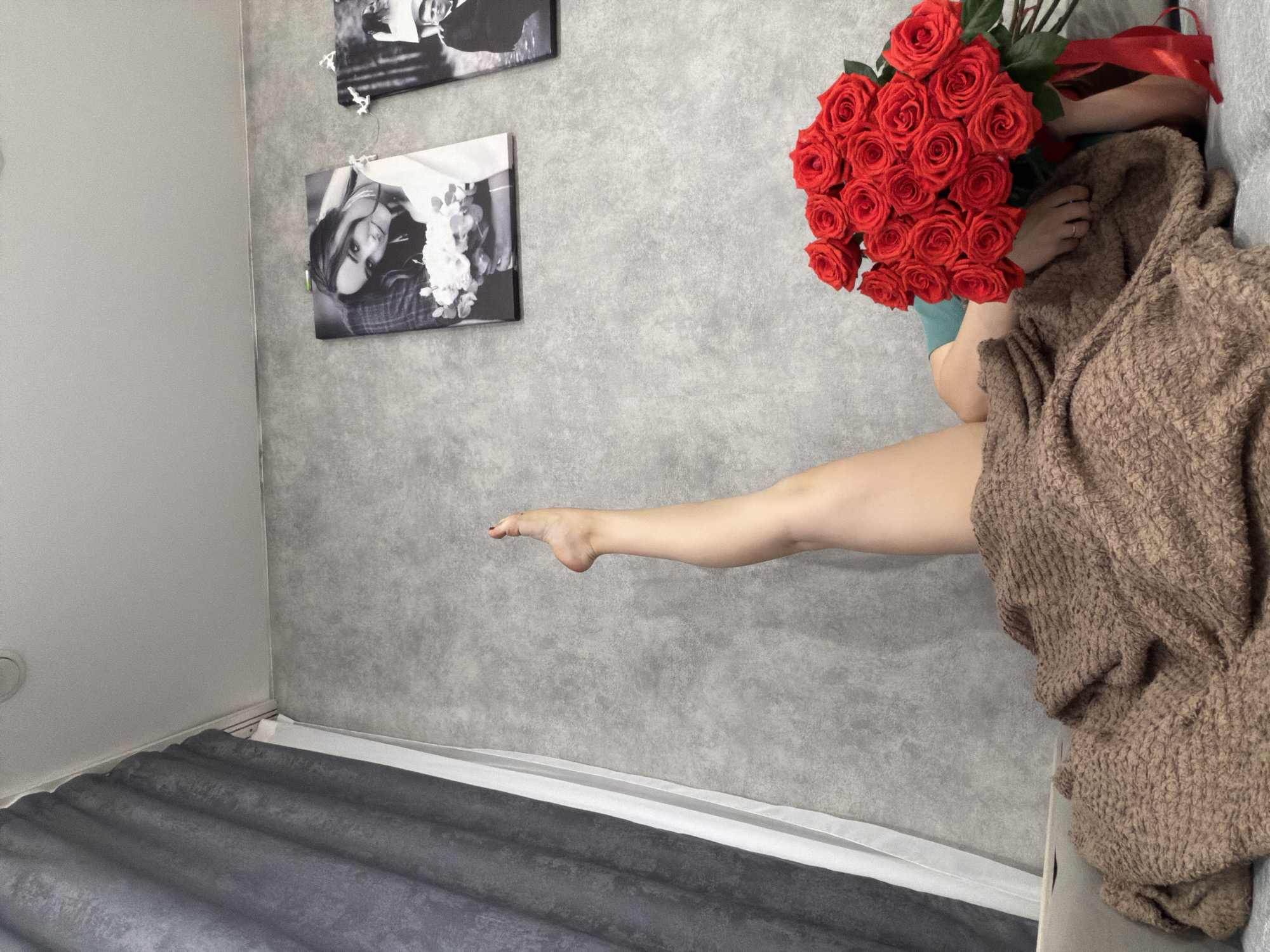 The image depicts an interior scene within a room. The room's walls are gray. Two black and white photographs hang on the wall. A bouquet of red roses is present. A person lies on the floor or a low surface. The person is partially covered with a brown knitted garment. One leg is extended. The location may be Chaikovsky, Russia, but this is not verifiable from the image.