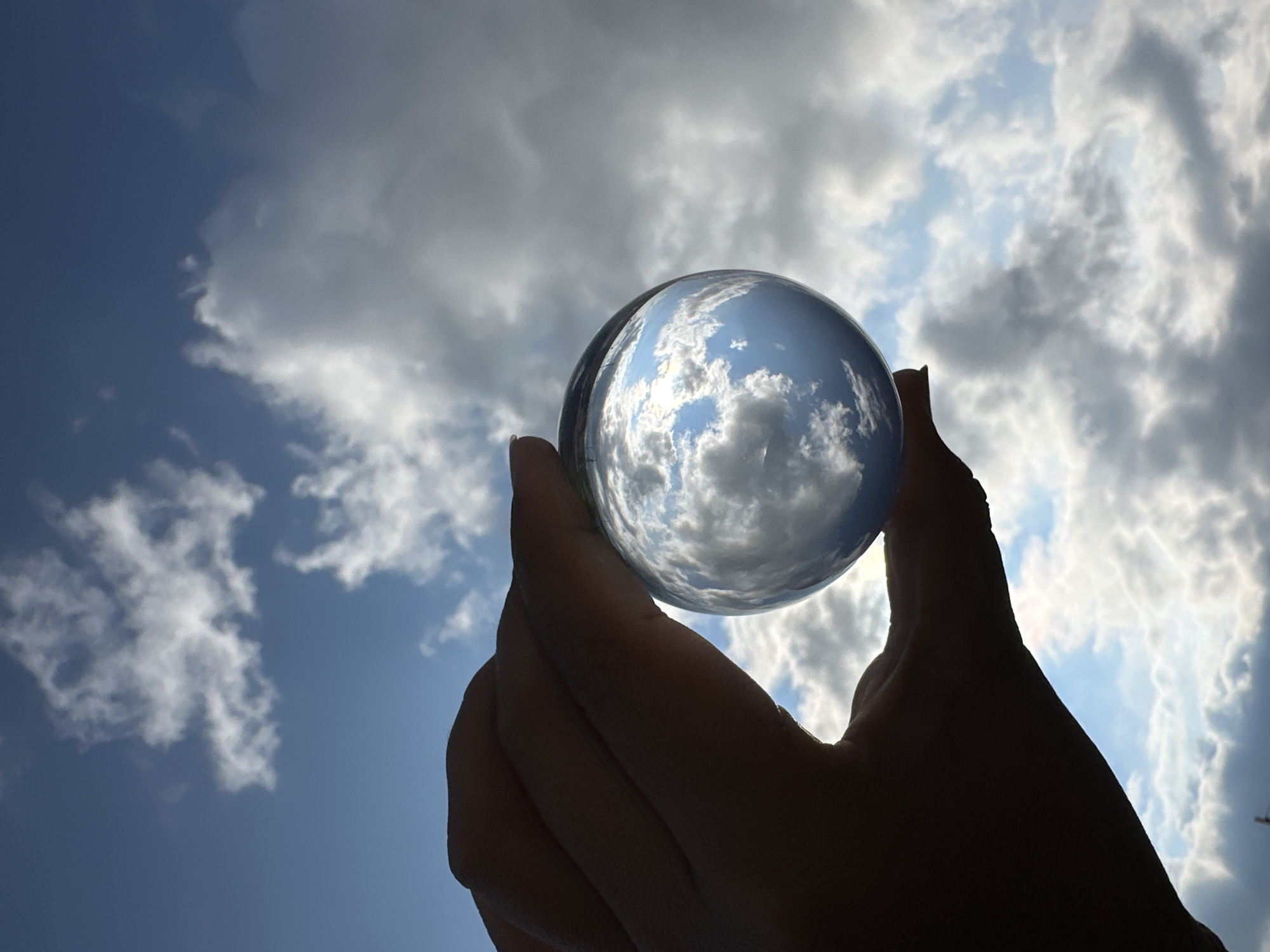 This media file features a close-up, low-angle shot of a human hand holding a clear glass sphere against a backdrop of a cloudy sky. The sphere, held between the thumb and index finger of the silhouetted hand, acts as a lens, capturing and inverting an image of the sky within its curvature. The sky itself is a dynamic mix of bright, sunlit clouds and darker, shadowed formations, suggesting a transition in weather or a partly cloudy day.
The lighting indicates it is daytime, likely late morning or afternoon, given the brightness of the sky and the contrast between the light and shadow in the clouds. The hand is silhouetted against the sky, obscuring details of skin tone or any jewelry. The focus is sharp on the glass sphere and the reflected sky, while the hand and the background sky are slightly softer.
The overall mood is one of contemplation or observation, with the sphere acting as a miniature world or a window into another perspective. The composition draws attention to the interaction between the human element and the natural environment, highlighting the reflective and refractive properties of the sphere. There is no visible text in the image. The setting is outdoors, with the sky being the dominant feature of the scene.