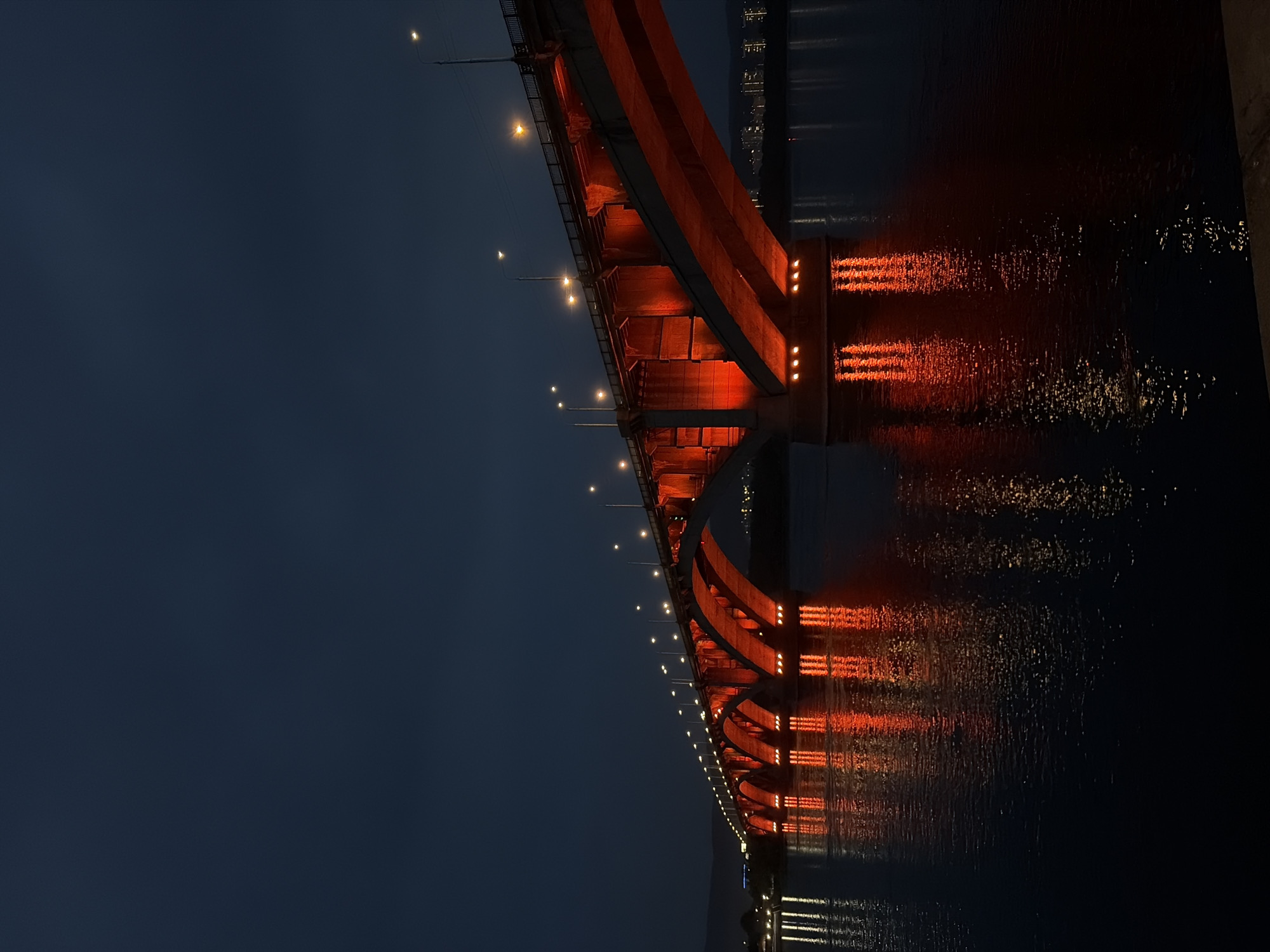 The image shows the Krasnoyarsk Railway Bridge, also known as the "Big Kazachinsky Bridge," spanning the Yenisei River in Krasnoyarsk, Russia. The bridge is illuminated with a vibrant red light, casting a strong reflection on the dark water below. The sky is a deep twilight blue, with a few distant lights visible on the far bank, suggesting a city setting. Several streetlights are also visible along the top of the bridge. The water is calm, reflecting the lights of the bridge and the sky. The perspective is from a low angle, looking up at the imposing structure. There are no people visible in the image.