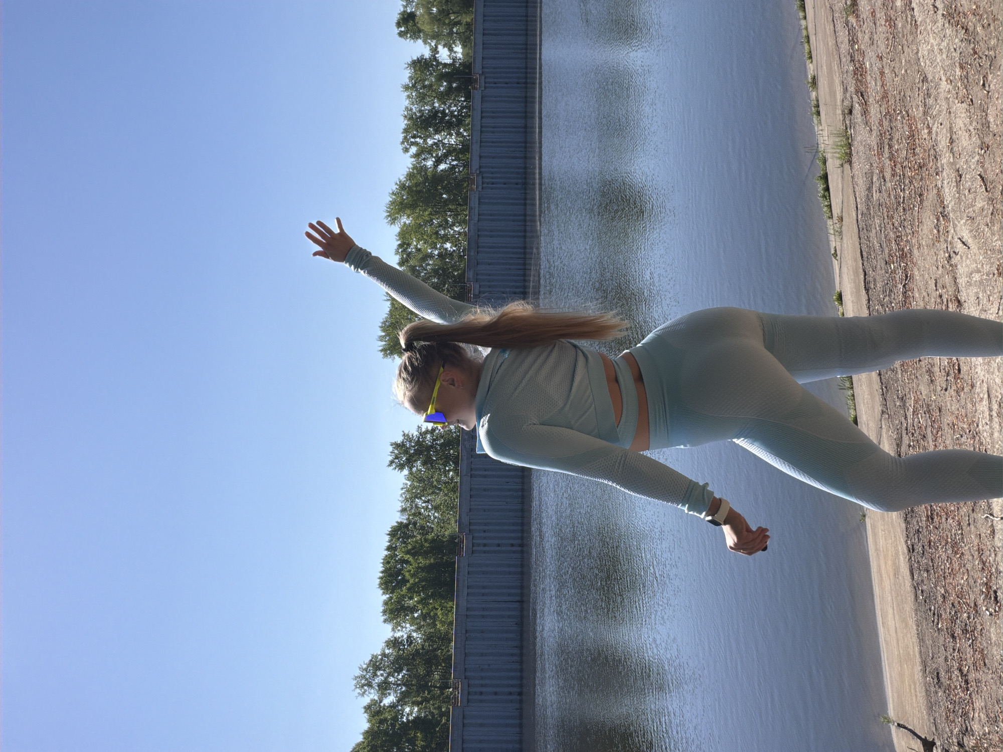 A woman in a light blue athletic outfit, including long-sleeved top, leggings, and sunglasses, is pictured in an outdoor setting in what appears to be the city of Chaikovsky, Russia, judging by the river and embankment structures typical of the area. She has her right arm raised towards the sky and her left arm is at her side, with a smartwatch visible on her wrist. She is standing near a body of water, possibly the Kama River, with trees lining the opposite bank and the sky visible in the upper left. The embankment is constructed of grey metal panels, and a concrete or paved path runs along the water's edge. The image has been rotated so that the horizon appears vertical.