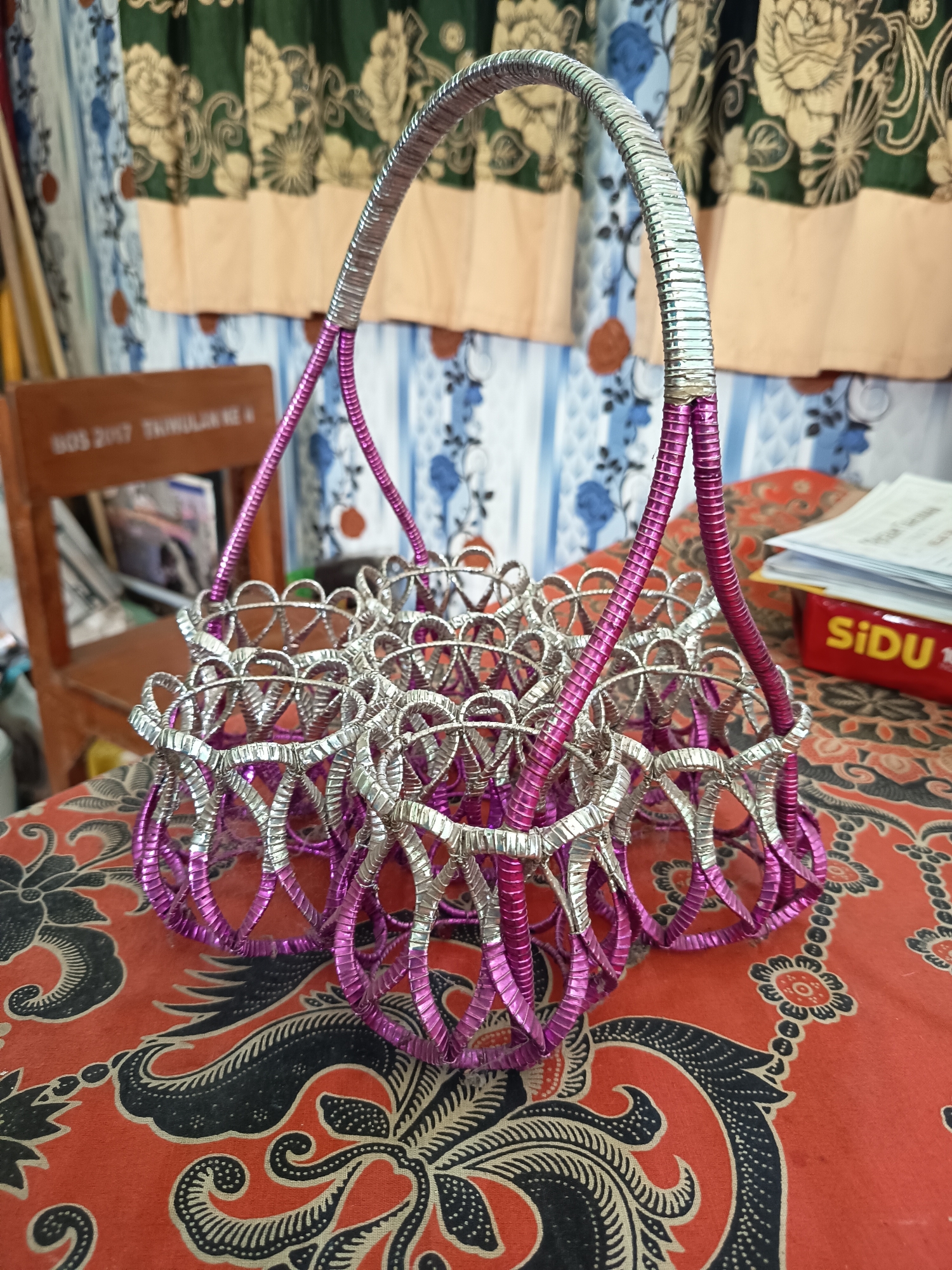 The image shows a handmade basket made of intertwined silver and purple metallic threads. The basket has a central handle formed by two strands of the metallic threads, which then split into smaller compartments at the base. The basket is positioned on a red tablecloth with a black batik pattern. In the background, a wooden chair with text visible on its back is slightly out of focus, as are patterned curtains and a red box with the text "SiDU" on it. The scene appears to be indoors, possibly in a home or a small shop. The lighting suggests it is daytime. There are no people visible in the image. The overall impression is that of a decorative item displayed in a domestic setting.