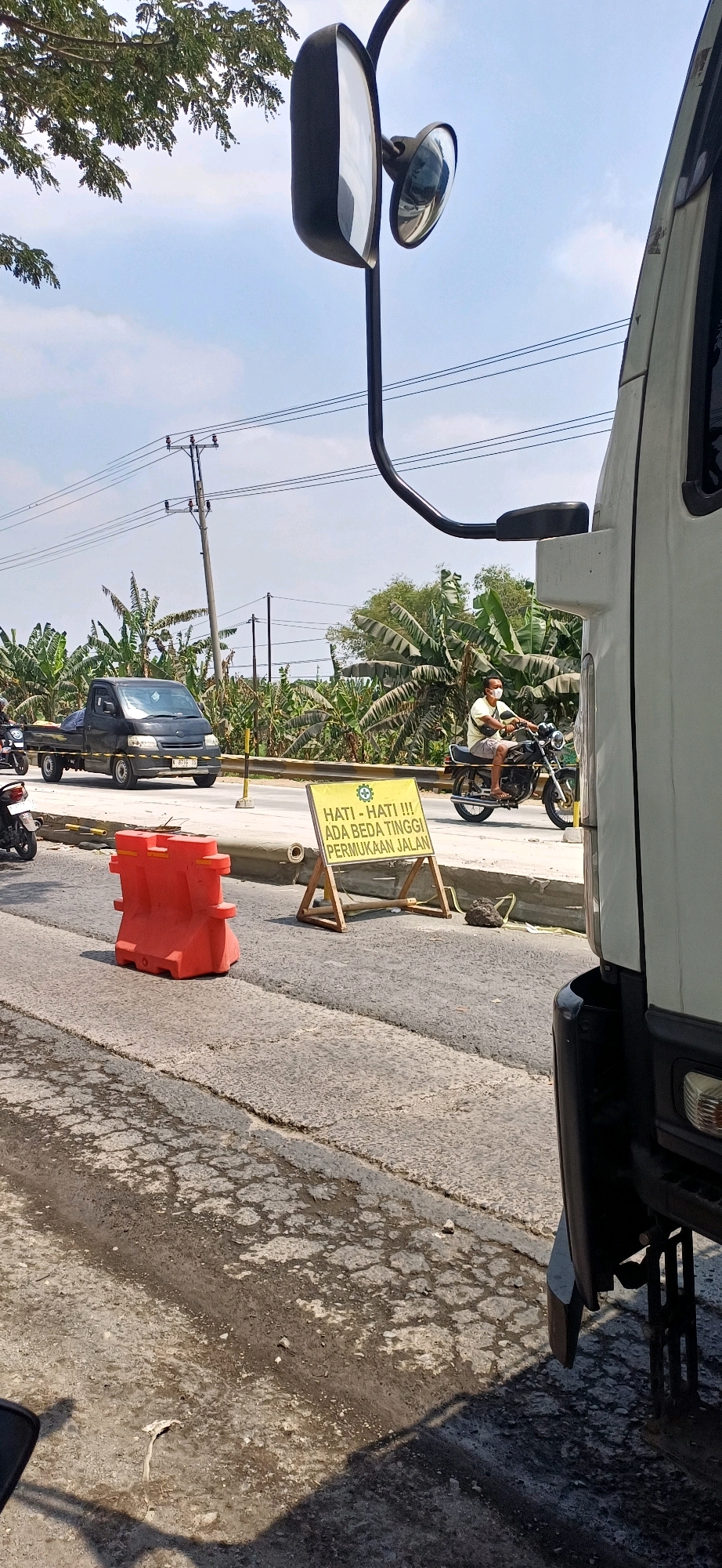 The image captures a street scene in Kecamatan Pati, Indonesia, on a sunny day. In the foreground, a portion of a white truck is visible on the right side. The road surface displays patches of wetness and cracked asphalt. To the left, an orange traffic barrier is placed on the road. Further back, a yellow sign with a green circular emblem and text in Indonesian is propped up on a wooden stand. The sign reads "HATI - HATI !!! ADA BEDATINGGI PERMUKAAN JALAN", which translates to "BE CAREFUL !!! THERE IS A HIGH ROAD SURFACE". A man wearing a light-colored t-shirt and shorts is riding a motorcycle, facing towards the right, with a light-colored mask on his face. Behind him, a black pickup truck is driving on the road. The background features lush green banana trees and utility poles with overhead power lines. The sky is clear and blue.