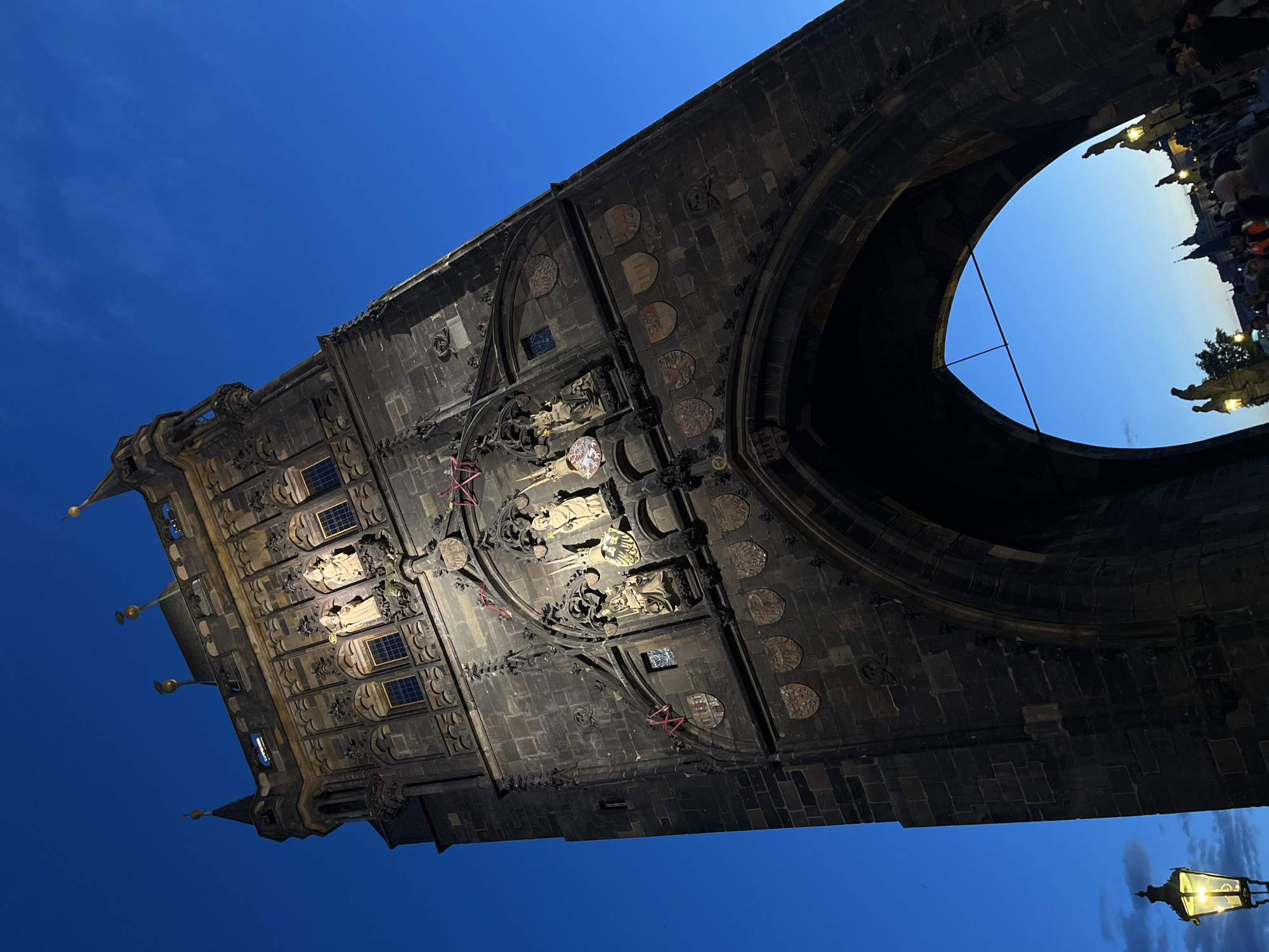 The image displays a grand, dark stone structure, likely a tower or an archway of a historic bridge or city gate, against a deep blue evening sky. The photo is taken from a low, angled perspective, looking upwards, with the structure dominating the frame.
The central subject is a massive piece of architecture, characterized by its intricate Gothic design. It is constructed from large, dark stone blocks and features multiple tiers of decorative elements. Prominent details include several statues housed in ornate niches, some adorned with what appear to be crowns or halos. Below these statues, numerous circular and shield-shaped emblems or coats of arms are embedded into the stone facade. Several small, arched windows are visible on the upper parts of the structure. Artificial lighting illuminates sections of the facade, highlighting the carvings and contributing to the nocturnal atmosphere. Some reddish-pink wires or cords are noticeable across parts of the stone surface.
Through the large, rounded archway, a lighter hue of blue sky is visible, along with silhouettes of numerous people gathered or walking. Distant architectural forms and illuminated statues or lampposts can also be faintly seen through the opening, suggesting a bustling public area beyond the arch.
The deep blue color of the sky, combined with the artificial lighting on the structure and a traditional street lamp glowing warmly in the bottom right corner, indicates that the scene takes place at dusk or night. The sky is clear, with minimal cloud cover. The ornate and ancient appearance of the stonework strongly suggests a historic European city setting. There is no readable text visible on the structure.