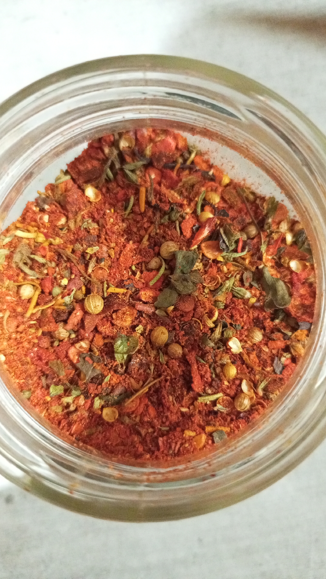The image shows a close-up, overhead view into a glass jar filled with a spice mix. The mix appears to be predominantly red, suggesting a base of chili powder or paprika, and also contains identifiable green herbs (possibly oregano, rosemary, or thyme), small round seeds (likely coriander), and other fragmented dried ingredients. The jar is clear, allowing the contents to be fully visible, and the rim is in focus. The background is a neutral, light-colored surface. The image doesn't provide information to confirm the location in Юдино, Russia.