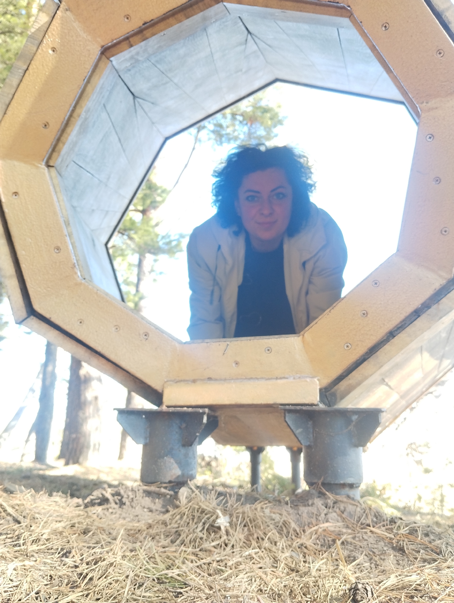 A woman with dark curly hair is looking through a hexagonal tunnel-like structure in a park-like setting. The structure is painted tan with black edges and is held up by four metal pillars. The ground is covered in dry grass and pine needles. Trees are visible in the background. The location is Votkinsk, Russia.