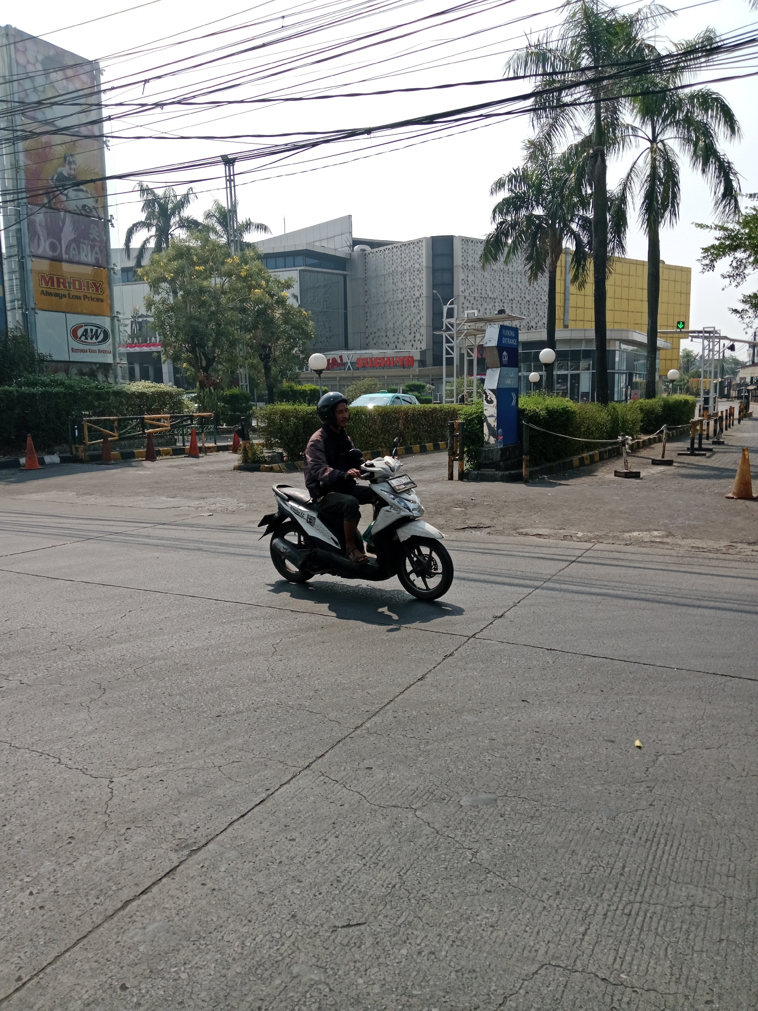An outdoor, eye-level, full shot shows a man riding a white scooter on a cracked asphalt road in front of a commercial building. The man is wearing a dark jacket and a black helmet. He is positioned in the center of the image, riding away from the camera. The building behind him is modern, with a yellow facade and white patterned walls. Palm trees are visible on the right side of the building. A large billboard with advertisements is on the left side of the building. Several traffic cones and barriers are placed along the side of the road. The sky is clear and bright. The location is identified as East Jakarta, Indonesia.