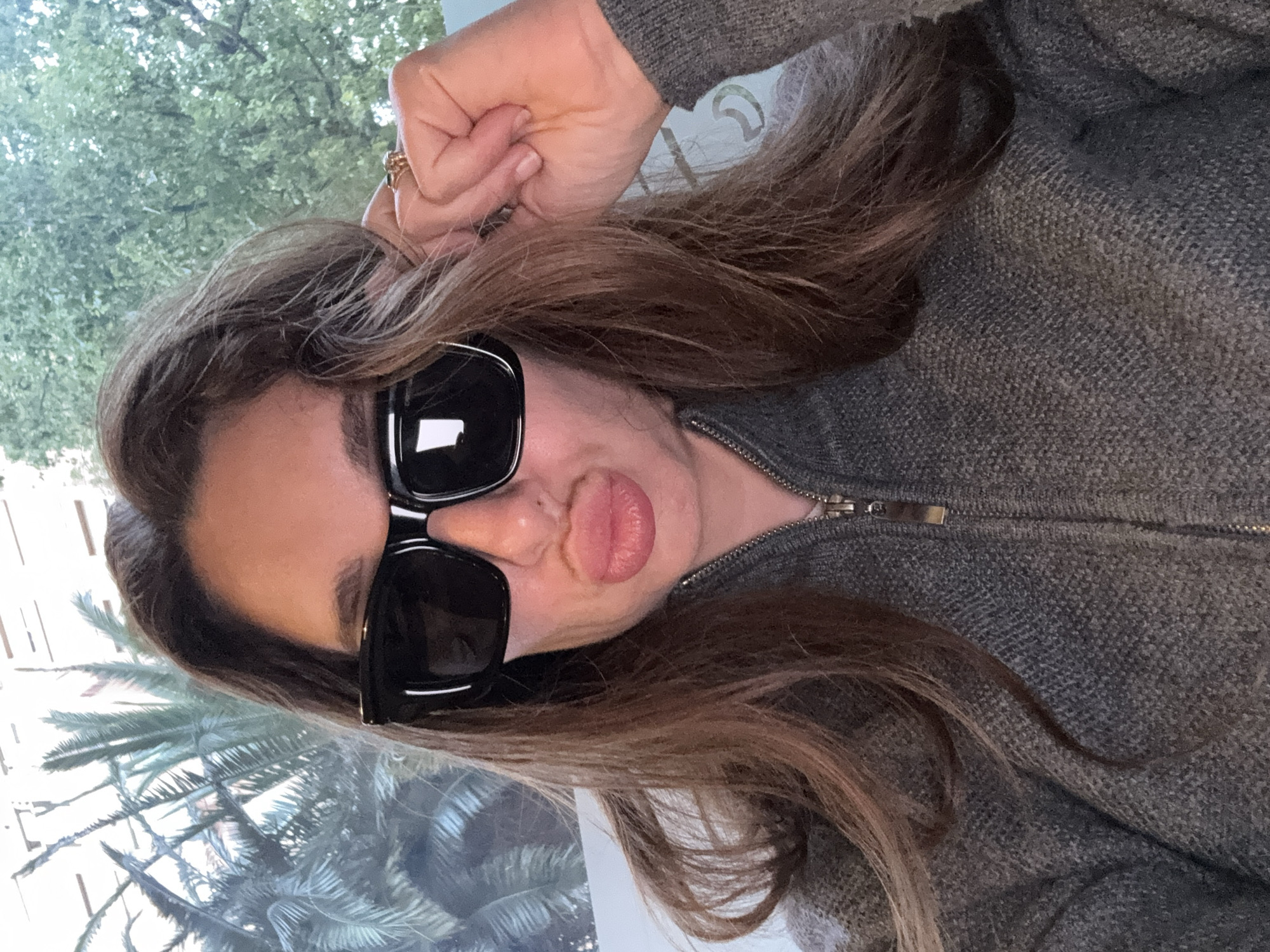 A medium close-up portrait, slightly tilted, features a woman looking directly at the camera, likely taking a selfie. She is wearing large, black, square-framed sunglasses that completely cover her eyes. Her lips are puckered in a "duck face" expression. Her long, brown hair, parted on the side, flows around her face and over her shoulders, resting on a grey, knitted, zippered sweater or jacket. Her right hand is raised, with fingers touching her temple or hair, and a ring is visible on one finger. The background appears to be an outdoor scene or a view through a window, dominated by vibrant green foliage, including discernible palm fronds, suggesting a warm and sunny location consistent with València, Spain. The bright lighting indicates it is daytime. There is no visible text in the image.