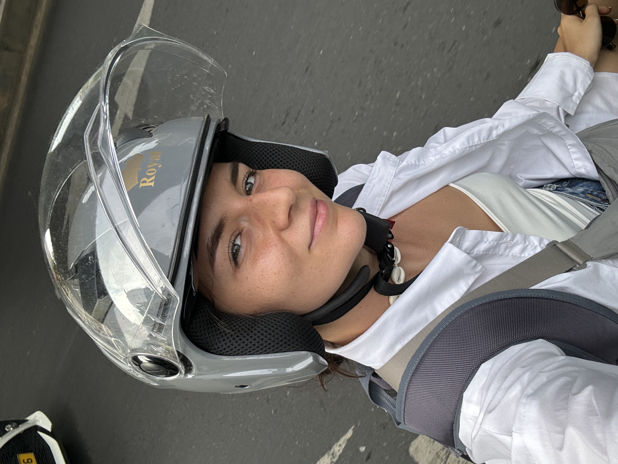 A medium shot, slightly angled, captures a young woman wearing a grey "Royal"-branded motorcycle helmet with a clear face shield. She's also wearing a white shirt and a white tank top. A backpack is strapped across her chest. She is looking directly at the camera. She is on a motorcycle in Thủ Đức, Vietnam.