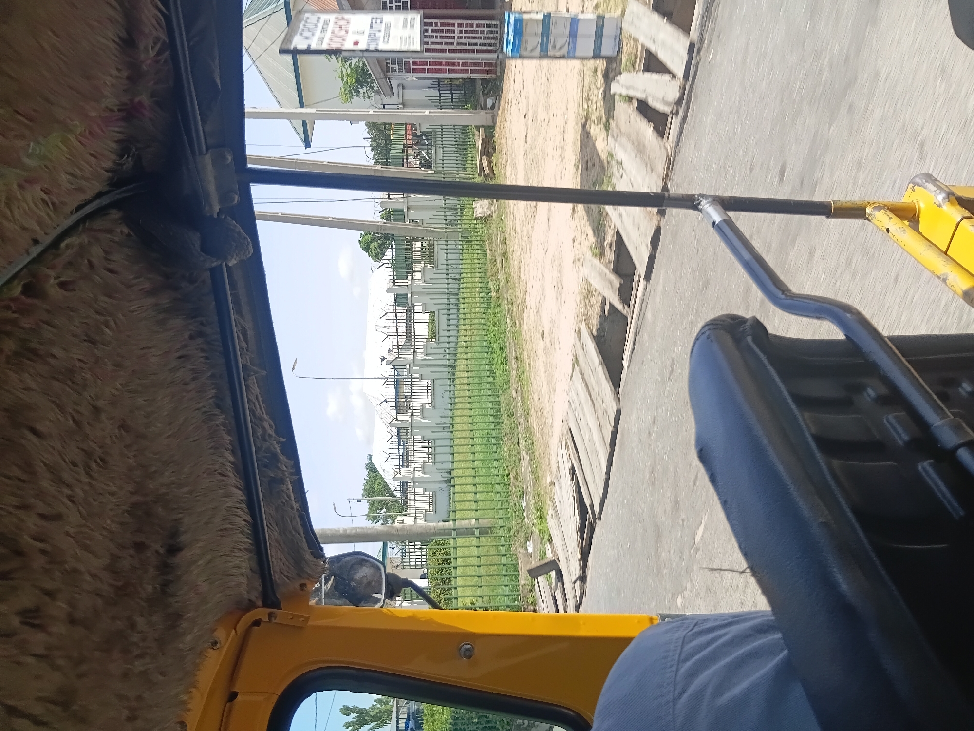 The image depicts a view from inside a yellow auto-rickshaw in Jalingo, Nigeria. The interior of the auto-rickshaw is lined with a textured, furry material on the ceiling. A person's shoulder, wearing a blue shirt, is visible in the foreground on the left. The auto-rickshaw is moving, as indicated by the blurred road and wooden planks forming a makeshift bridge over a ditch.
Beyond the auto-rickshaw, a green metal fence encloses a grassy area. Behind the fence, a white building with a sloped roof is visible, along with what appears to be a security fence with barbed wire on top. Several tall poles, possibly utility poles or fence supports, are also present. To the right of the building, a smaller structure with a red and white sign is visible. The sign has text, some of which is legible and reads "MOROCC... BOOKSHOP COMPUT...". Stacked blue and white containers are positioned near the sign. The sky is clear and blue. The overall scene suggests a street-level view within the city, likely near a commercial or institutional area.