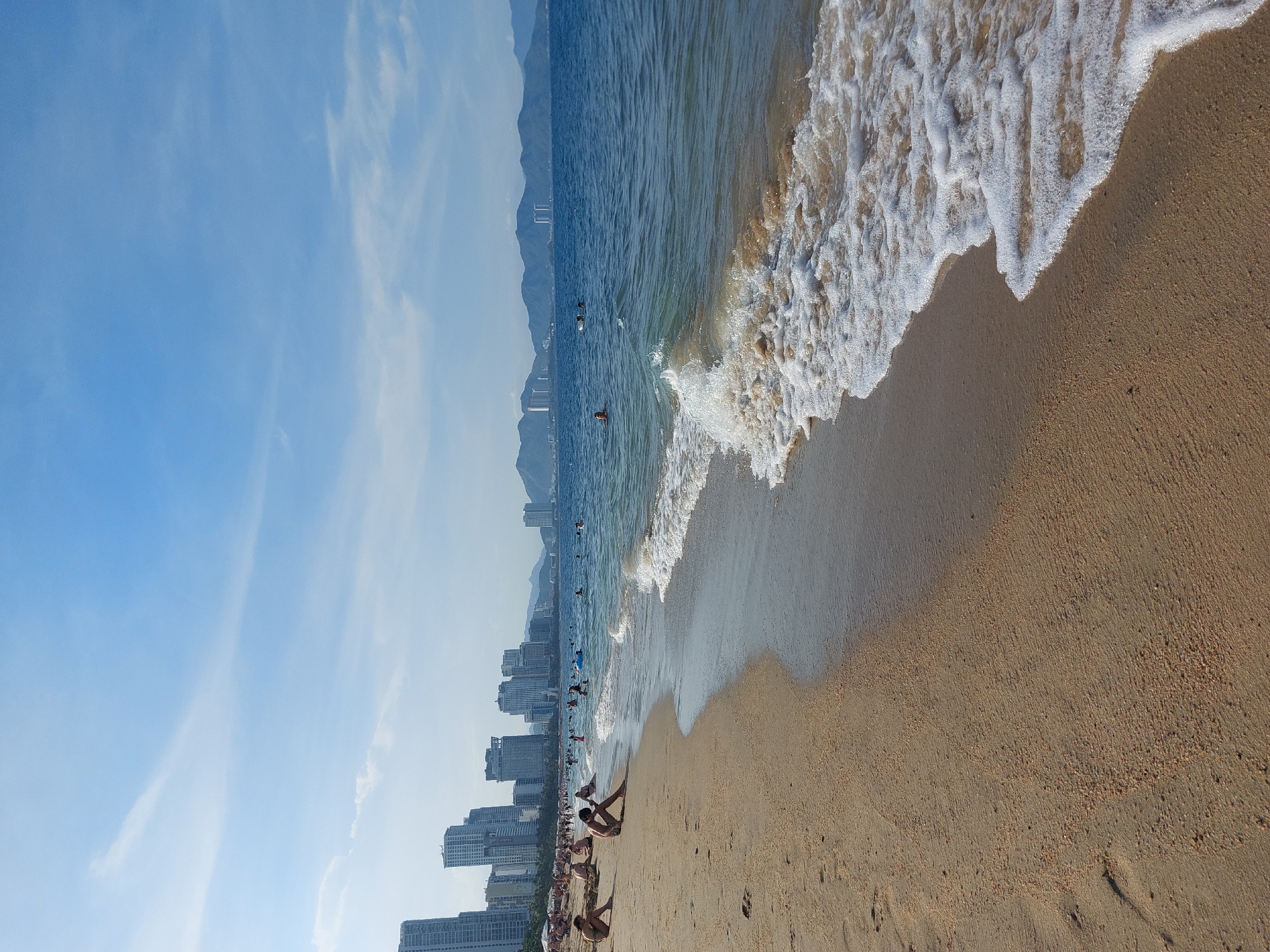 The image depicts a wide, sandy beach in Nha Trang Ward, Vietnam, with the azure sea meeting the shore. In the distance, a dense collection of high-rise buildings lines the coastline, characteristic of a modern city. The sky above is a clear blue, with a few wispy white clouds.
On the beach, several individuals are visible. Some are seated on the sand, appearing relaxed. Others are in the shallow water, swimming or wading. A gentle wave with foamy crests is approaching the shore, adding a dynamic element to the scene. The sand itself is light brown and appears somewhat textured, with small particles and some footprints visible. The overall impression is of a popular, bustling beach on a sunny day in an urban coastal environment.