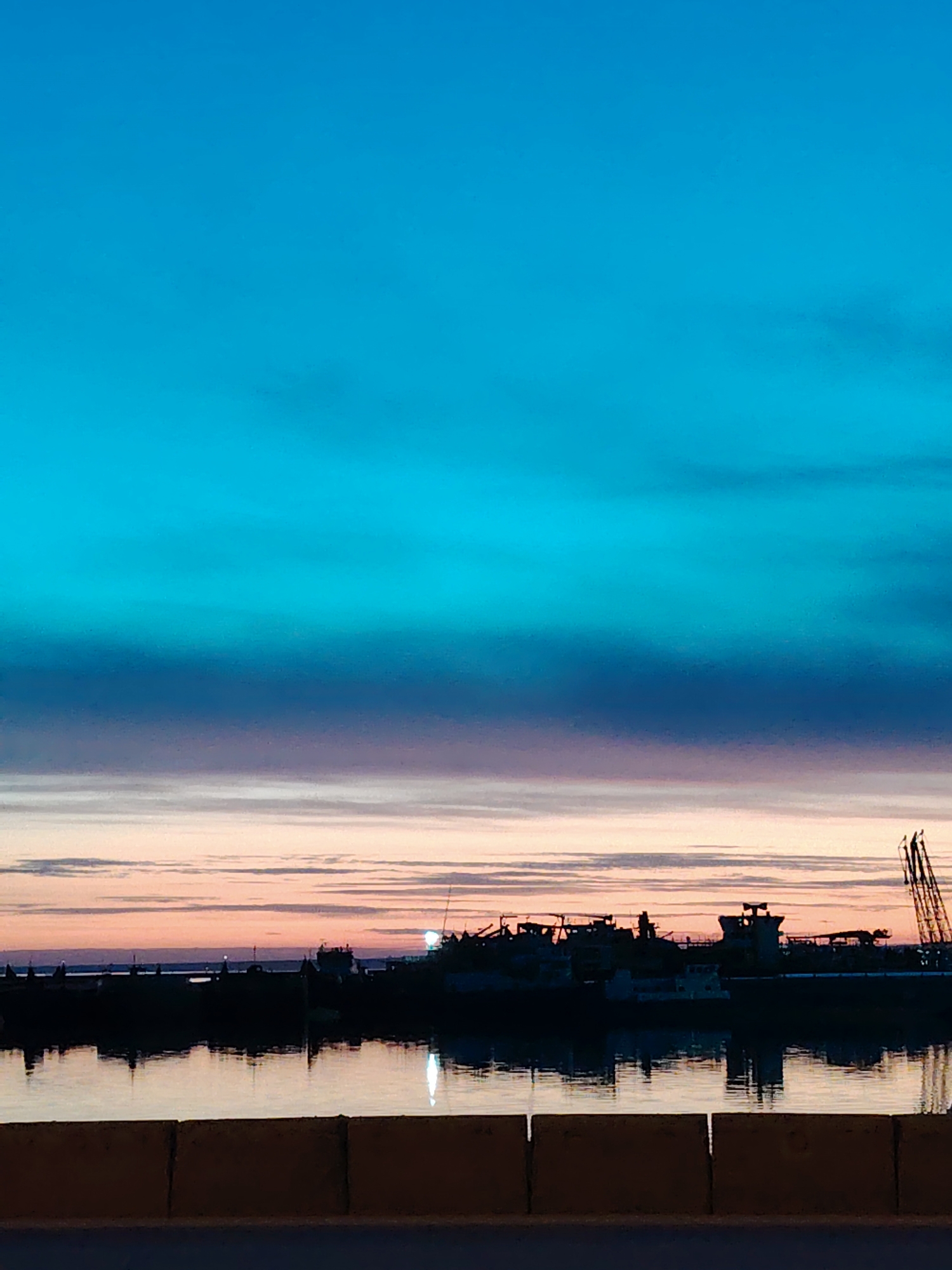 The image depicts a waterfront scene at dusk in Chaikovsky, Russia. The sky transitions from a vibrant turquoise at the top to layers of blue, lavender, and peach hues near the horizon. A silhouetted cityscape, possibly a port or industrial area, lines the water's edge. Structures resembling ships, cranes, and buildings are visible against the colorful sky. The water reflects the silhouette and the dim light, creating a mirrored effect. A low, yellow retaining wall or barrier is in the foreground. There are no people visible in the image.