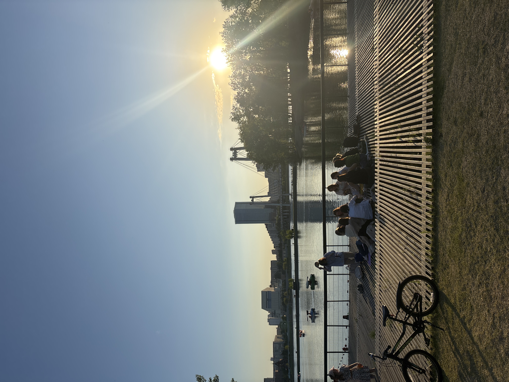 This media file captures a serene late afternoon or early evening scene along an urban waterfront, likely in Krasnoyarsk, Russia. The sun, low on the horizon, radiates a golden light and prominent lens flares from the upper left, casting long shadows and backlighting much of the scene.
In the foreground, a modern wooden deck or walkway with parallel slats stretches along the right side of the frame. Several individuals are seated on this deck, silhouetted against the water or partially lit by the ambient glow, appearing to relax and enjoy the pleasant weather. One person stands by the railing, looking towards the water, while another is casually reclined on the deck further in the foreground. To the immediate right of the deck, a patch of green grass is visible, where a black bicycle is parked, leaning on its kickstand.
The deck borders a wide body of water, possibly the Yenisei River, whose surface reflects the bright sunlight. In the midground, several small recreational boats, resembling pedal boats, are visible on the water, suggesting leisure activities.
Across the water, a cityscape unfolds under a clear, blue sky. A prominent tall, modern building stands out among other urban structures. Further in the distance, a large suspension or cable-stayed bridge spans the water, connecting different parts of the city. Trees line the far bank, adding a touch of nature to the urban backdrop.
The overall atmosphere is one of peaceful relaxation and enjoyment of the evening sun in an urban riverside setting. No discernible text is visible within the image.