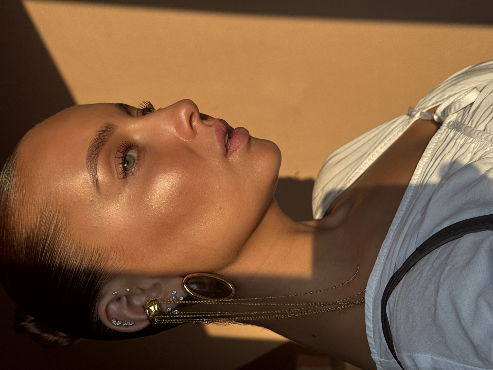 This is a close-up, profile shot of a young woman from the chest up, looking to the right. She has her dark hair pulled back tightly into a bun. Her skin is illuminated by golden hour sunlight, highlighting her features. She has several piercings in her left ear, including cartilage piercings adorned with small gems and a statement earring featuring multiple gold chains and a large, dark oval stone. She is wearing a white top with a textured fabric and a dark strap from a bag is visible across her shoulder. The background is a warm, orange wall with a distinct shadow line on the right side. The scene suggests an outdoor setting during sunset. There is no information or visual cue that places this image in Chaikovsky, Russia.
