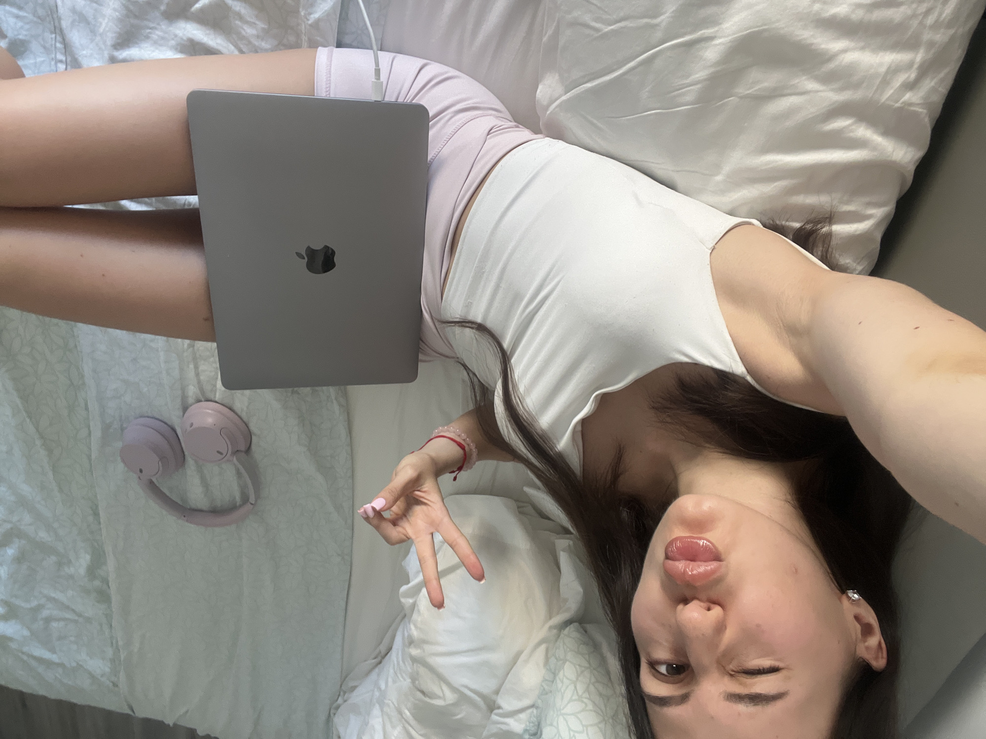 The image shows a young woman lying on a bed with a laptop on her legs. She is wearing a white tank top and pink shorts. A pair of pink headphones are placed on the bed next to her. The woman is looking at the camera and making a peace sign with her hand. She has long dark hair and is wearing lip gloss. She is also wearing a red bracelet on her left wrist and a diamond stud earring in her right ear. The room appears to be a bedroom, with white bedding and pillows. The lighting suggests it is daytime. The context provided indicates the location is Palma de Mallorca, Spain.