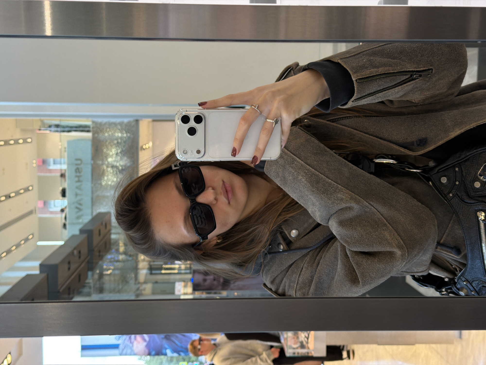 A person takes a selfie in a mirror. The person is wearing sunglasses and a gray jacket. A black purse hangs on their shoulder. The reflection in the mirror shows the interior of a building with the name "ЦУМ" (TSUM) visible, indicating the Central Universal Department Store in Moscow, Russia. A man is reflected in the bottom of the mirror. The person is holding a white smartphone with multiple camera lenses. They have rings on their fingers and short burgundy nail polish.
