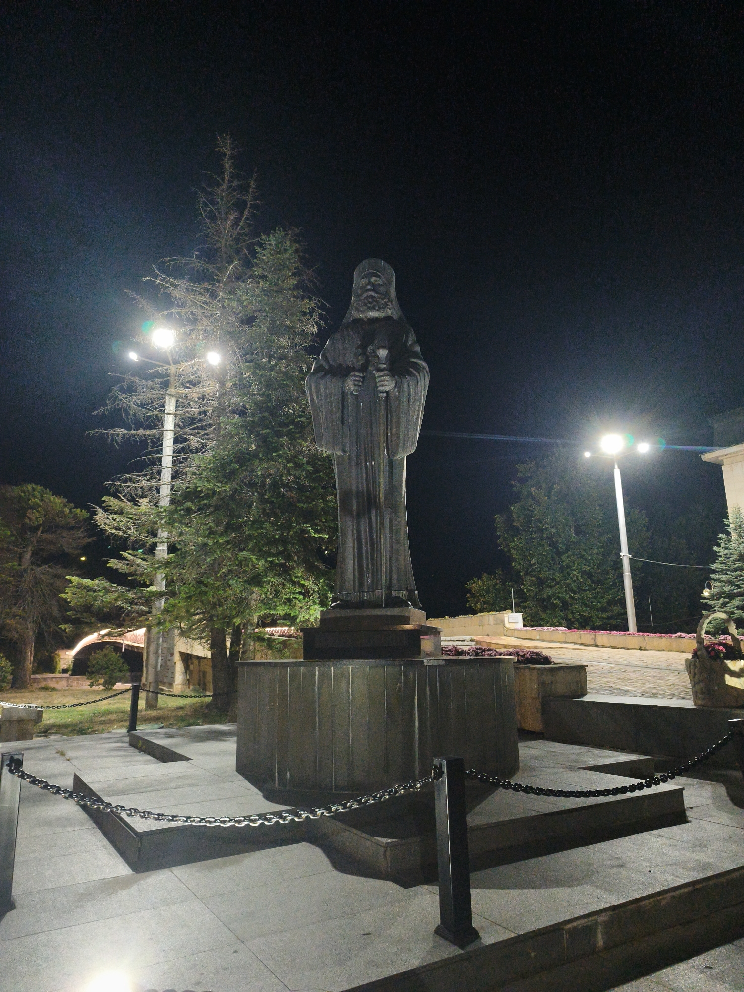 The image shows a night-time view of a large stone monument of a robed male figure holding a staff. The monument stands on a multi-tiered stone pedestal, surrounded by paved ground and low-lying decorative chains supported by posts. Behind the monument, trees and streetlights are visible. In the background, a bridge with an arched structure can be seen, suggesting an urban setting. The monument is located in the city of Lovech, Bulgaria.